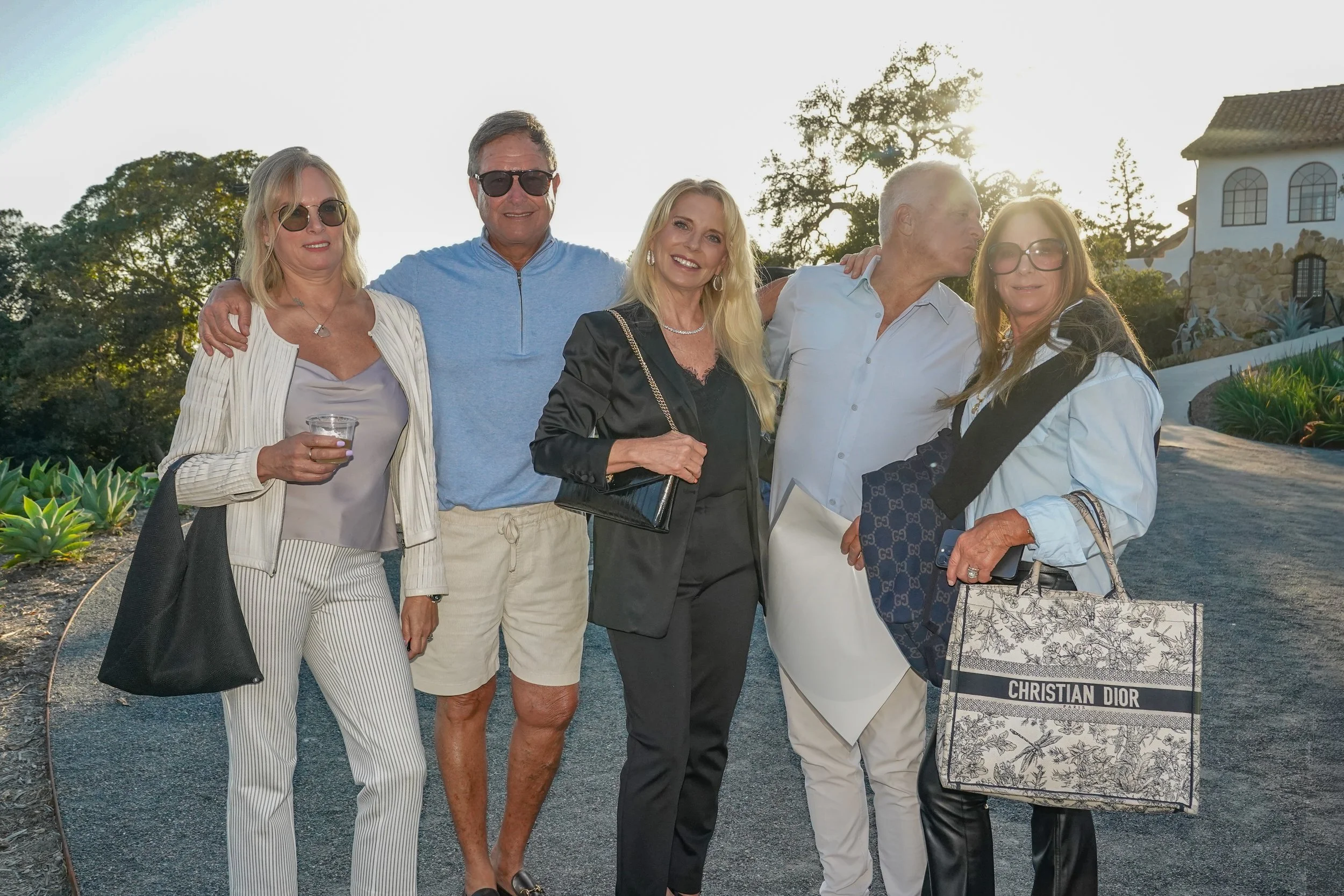 Group of six adults posing outdoors during sunset, dressed in casual and semi-formal clothing, with greenery and a white building in the background.