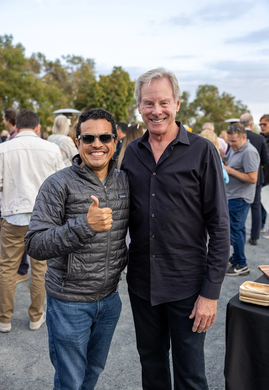 Two men smiling and posing for a photo outdoors, with one giving a thumbs-up. The man on the left wears sunglasses and a black quilted jacket, and the man on the right wears a black button-up shirt. There are people gathered in the background at what