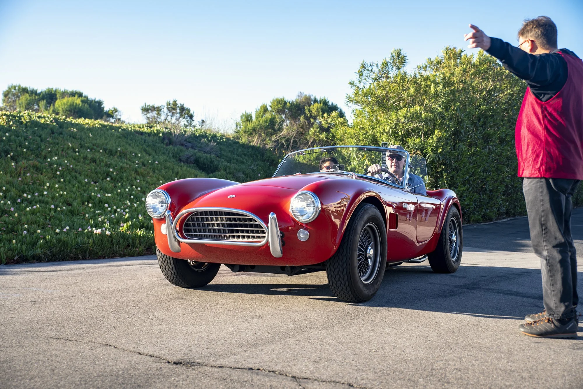 A red vintage convertible sports car with two people inside, one driving and one in the passenger seat, on a sunny day with a person standing outside the car gesturing towards it.