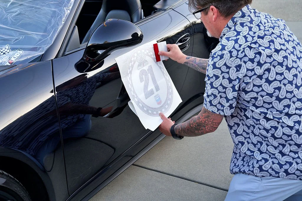 A man with tattoos on his arm and wearing glasses and a paisley-patterned shirt is applying a decal with the number 21 to a black car's side panel.