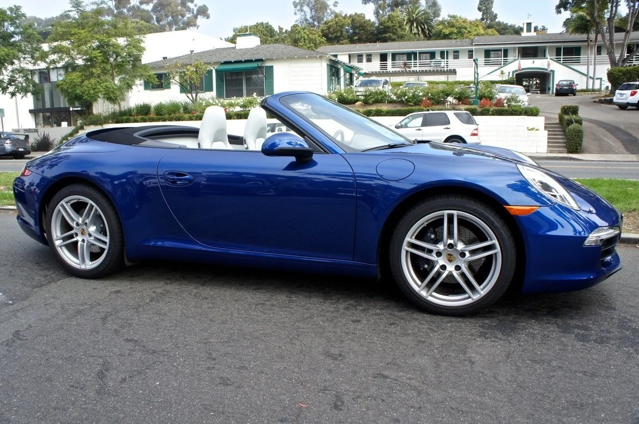 A blue convertible sports car parked on a city street with residential buildings and trees in the background.