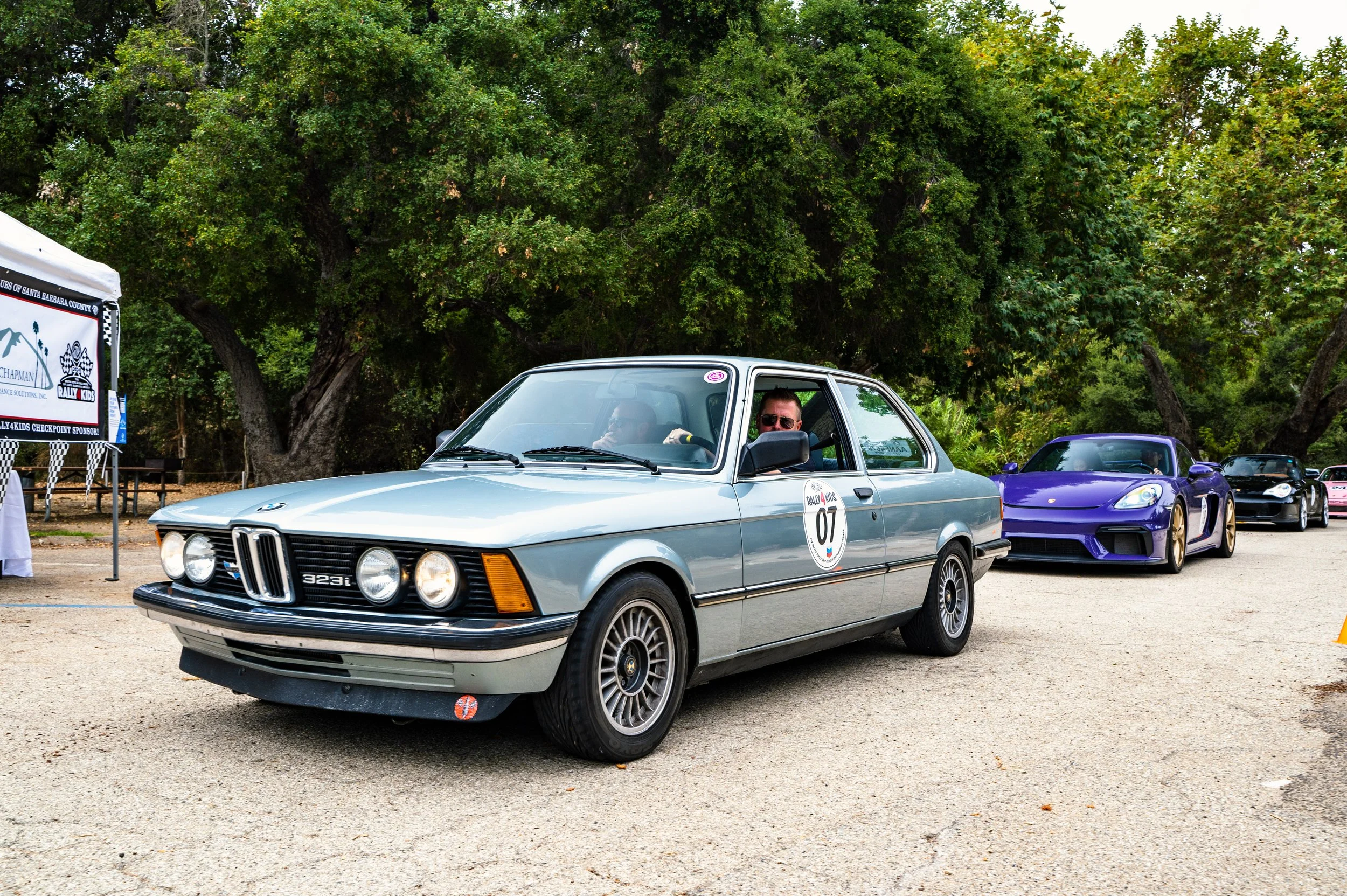 A vintage light blue BMW 3 Series E21 car with the number 07 on a rally sticker, parked on a gravel path during a car show. Behind it, a purple sports car and other cars are visible, with large green trees in the background.