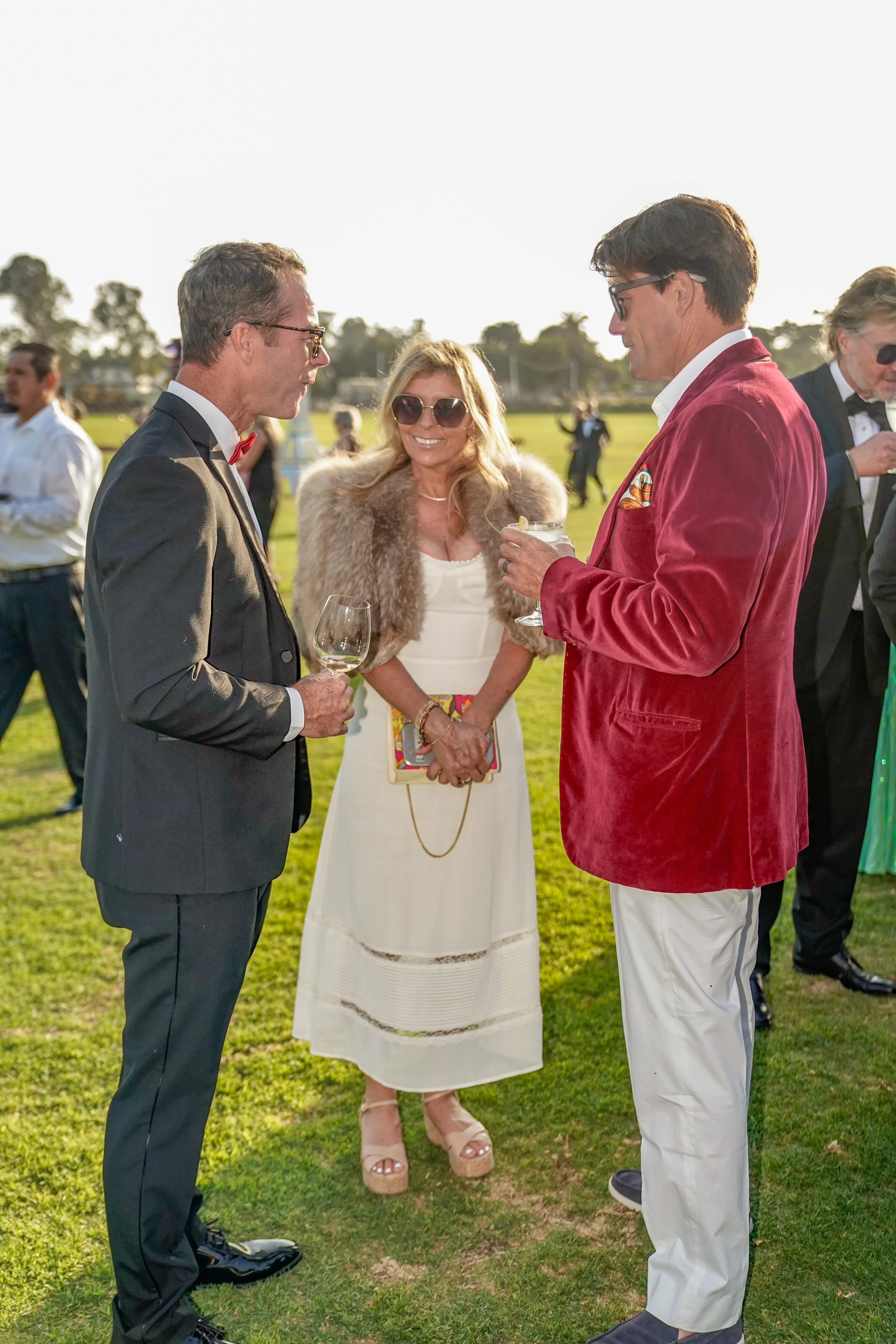 People in formal attire socializing outdoors, holding drinks, at a garden party or wedding reception.