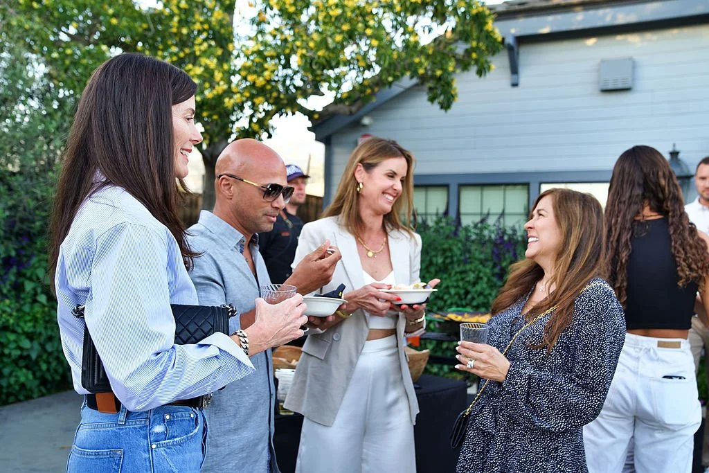 Group of people socializing outdoors, holding drinks and bowls, in a backyard with trees and a house in the background.