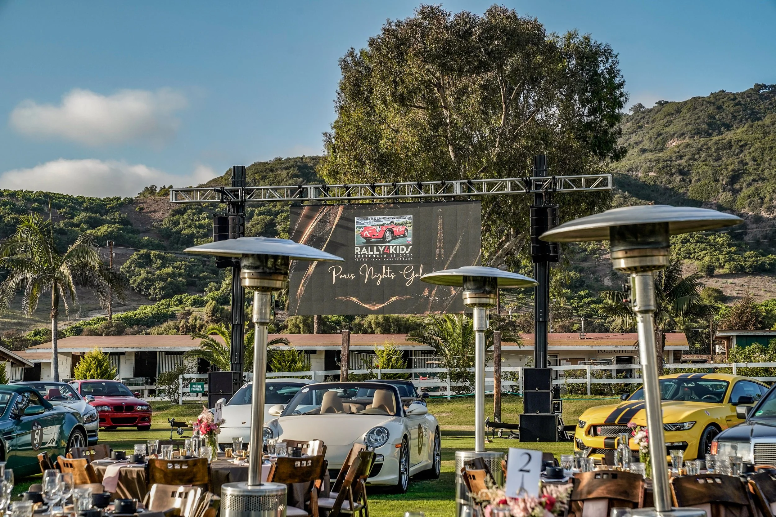 Outdoor event with luxury sports cars, banquet tables with chairs, and a large screen displaying a red car with the text 'RALLY4KID SEP 12 2025 PARIS NIGHTS GALA' in a scenic setting with green hills and palm trees.