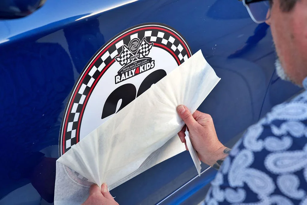 Person applying a decal sticker to a blue race car, with a Rally Kids logo and checkered flag design.