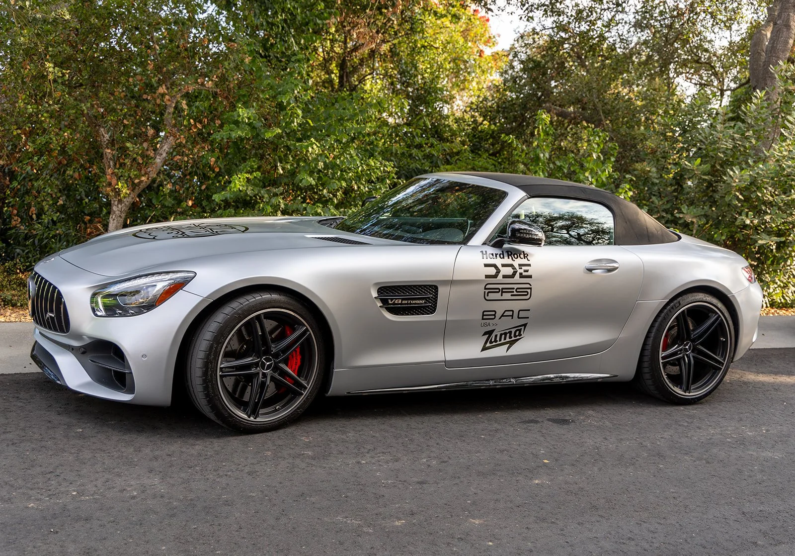Silver convertible car with black rims and red brake calipers parked on a road, with green trees and foliage in the background.