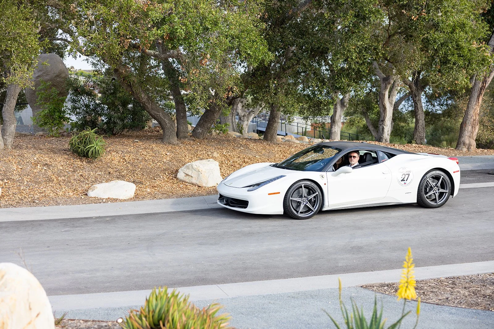A white sports car with a black roof, marked with the number 27, parked on a street with trees and rocks in the background, and a man with sunglasses inside.
