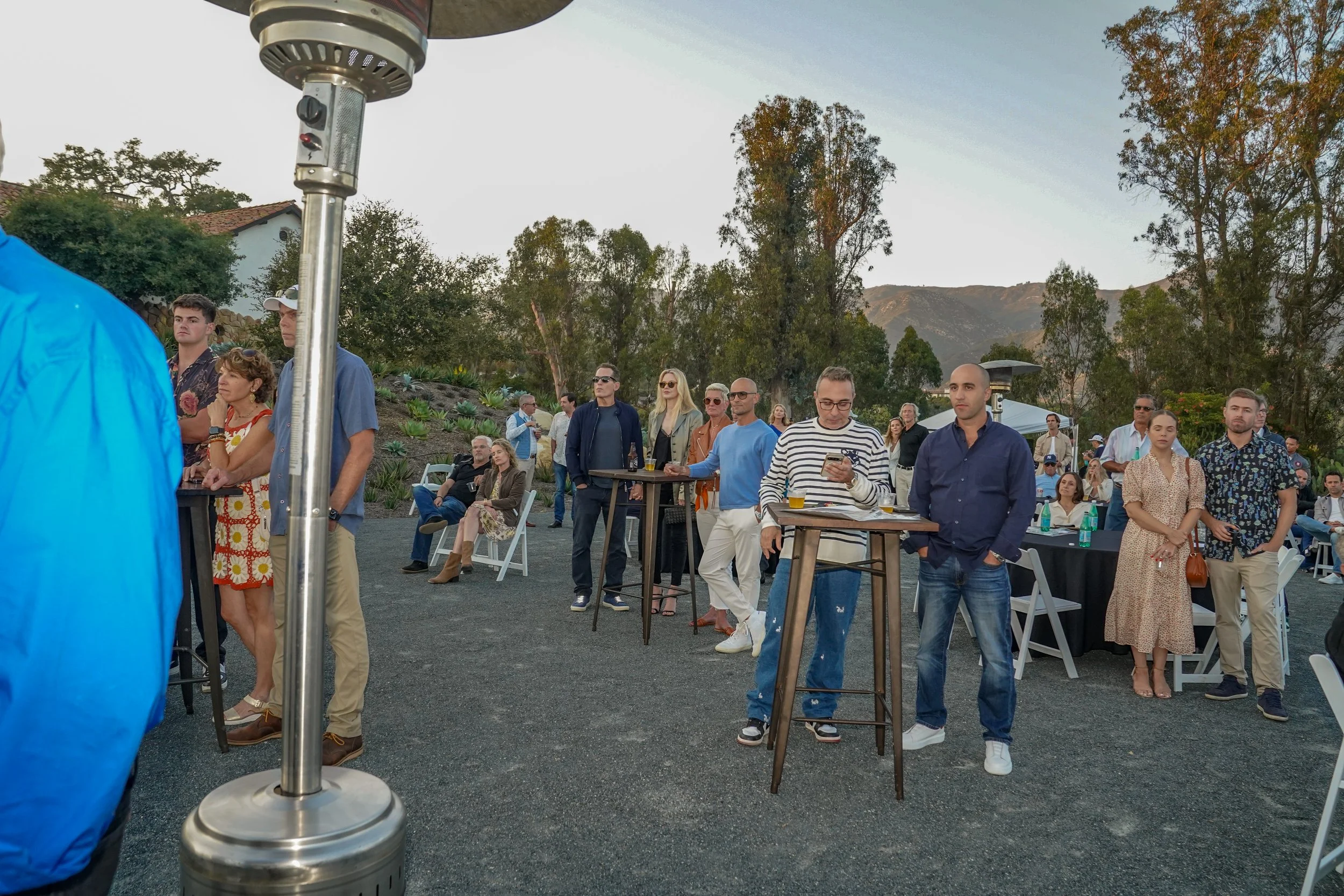 People attending an outdoor event, some standing and some sitting at tables, with trees and mountains in the background during sunset.