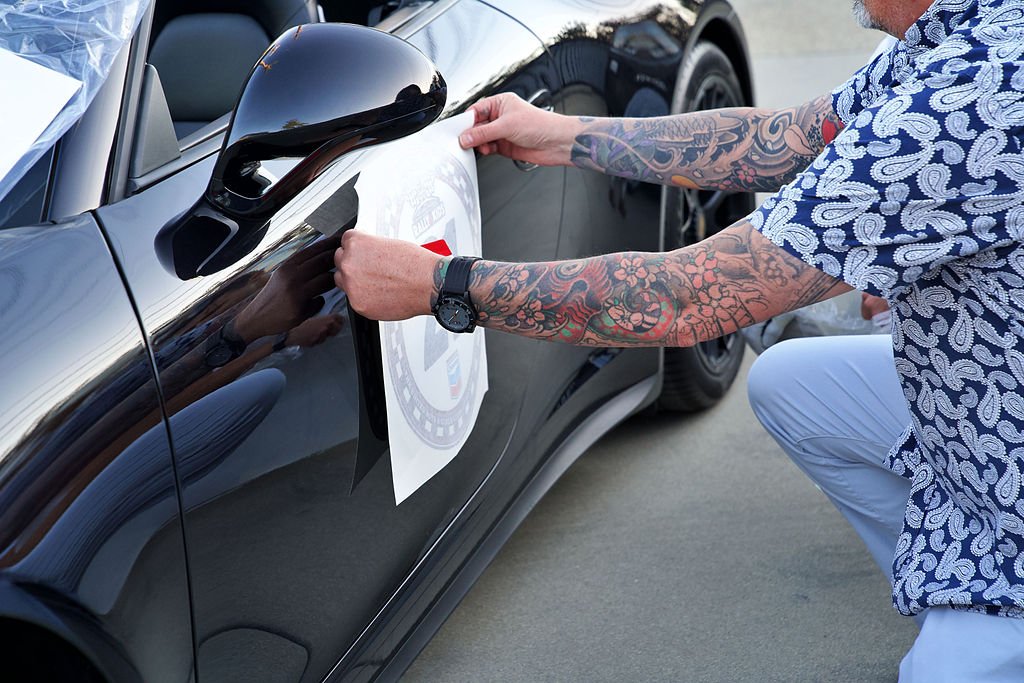 Person applying a decal or sticker to the side of a black sports car with a skull tattooed arm, wearing a patterned shirt.