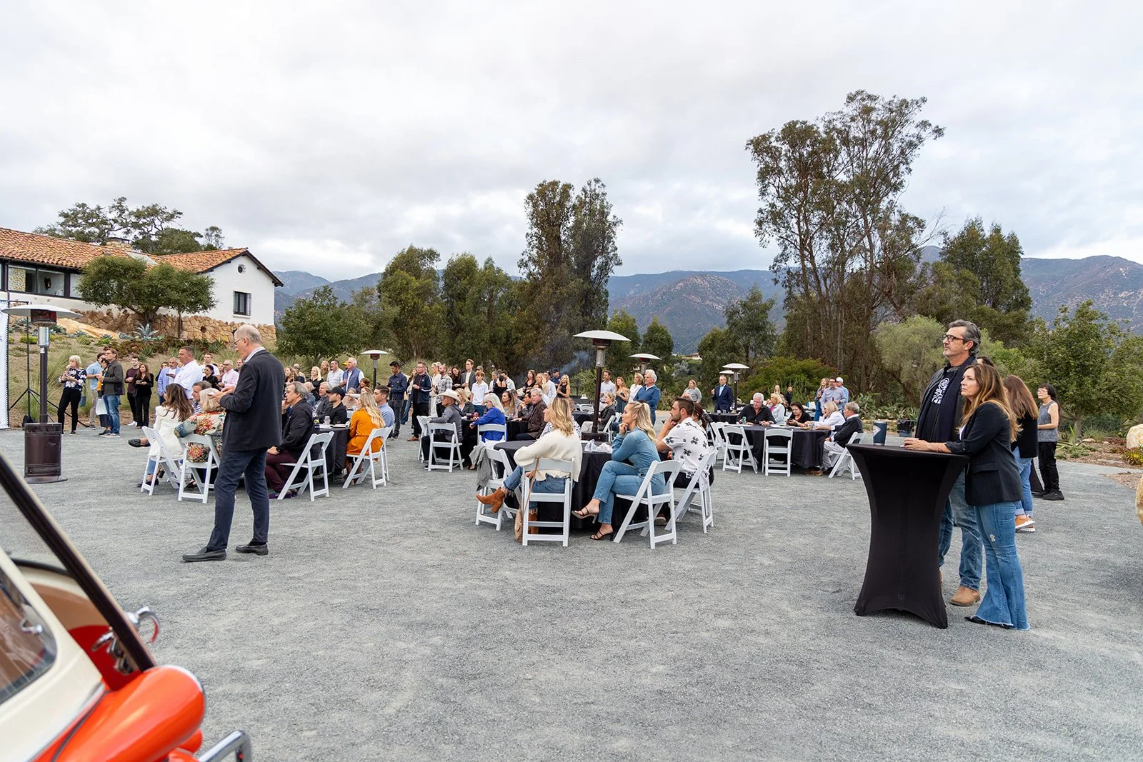 Outdoor gathering with people seated at tables and standing around on a gravel surface, with trees, mountains, and partly cloudy sky in the background.