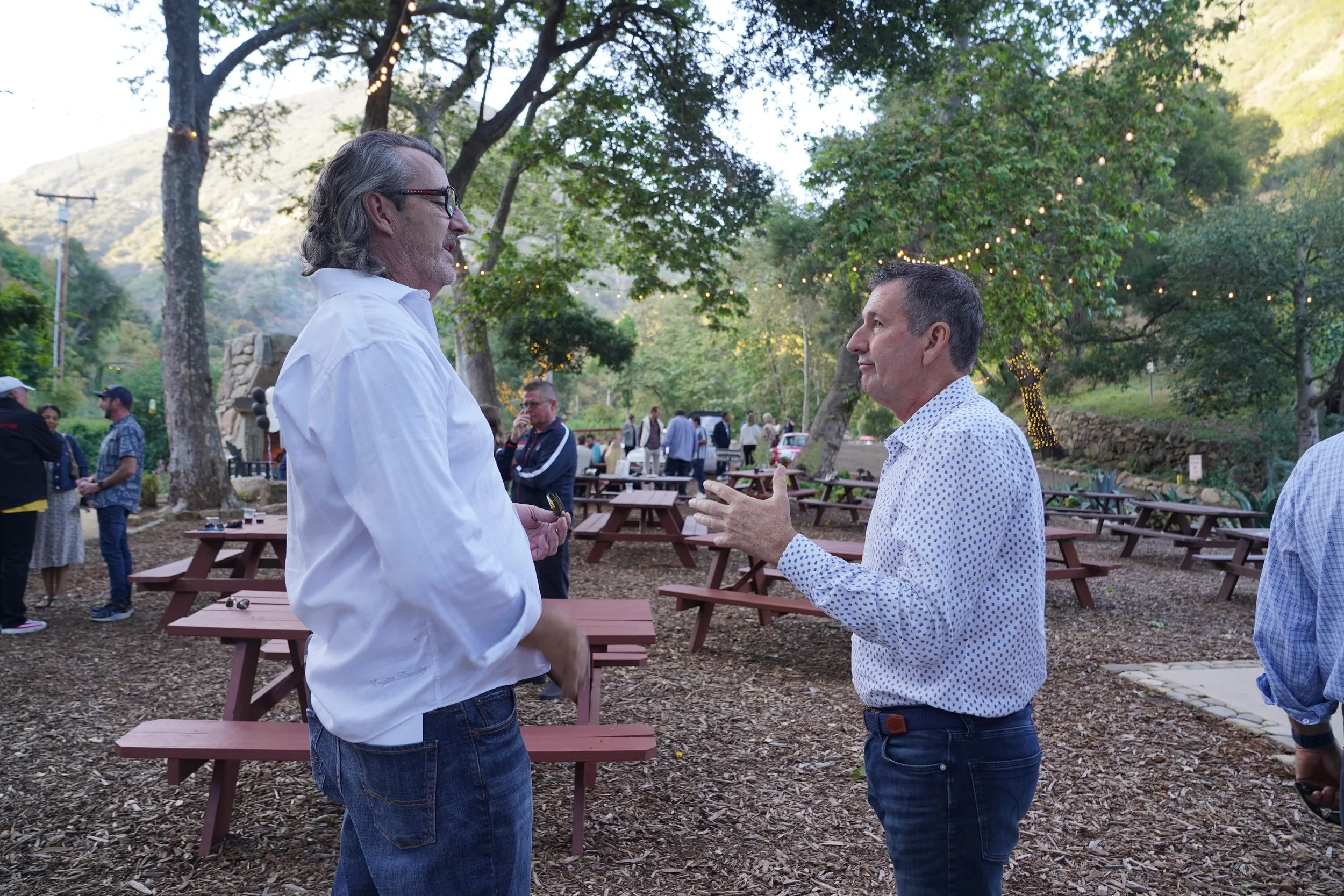 Two men are standing and talking outdoors in a park-like setting with picnic tables and string lights, surrounded by trees and hills.