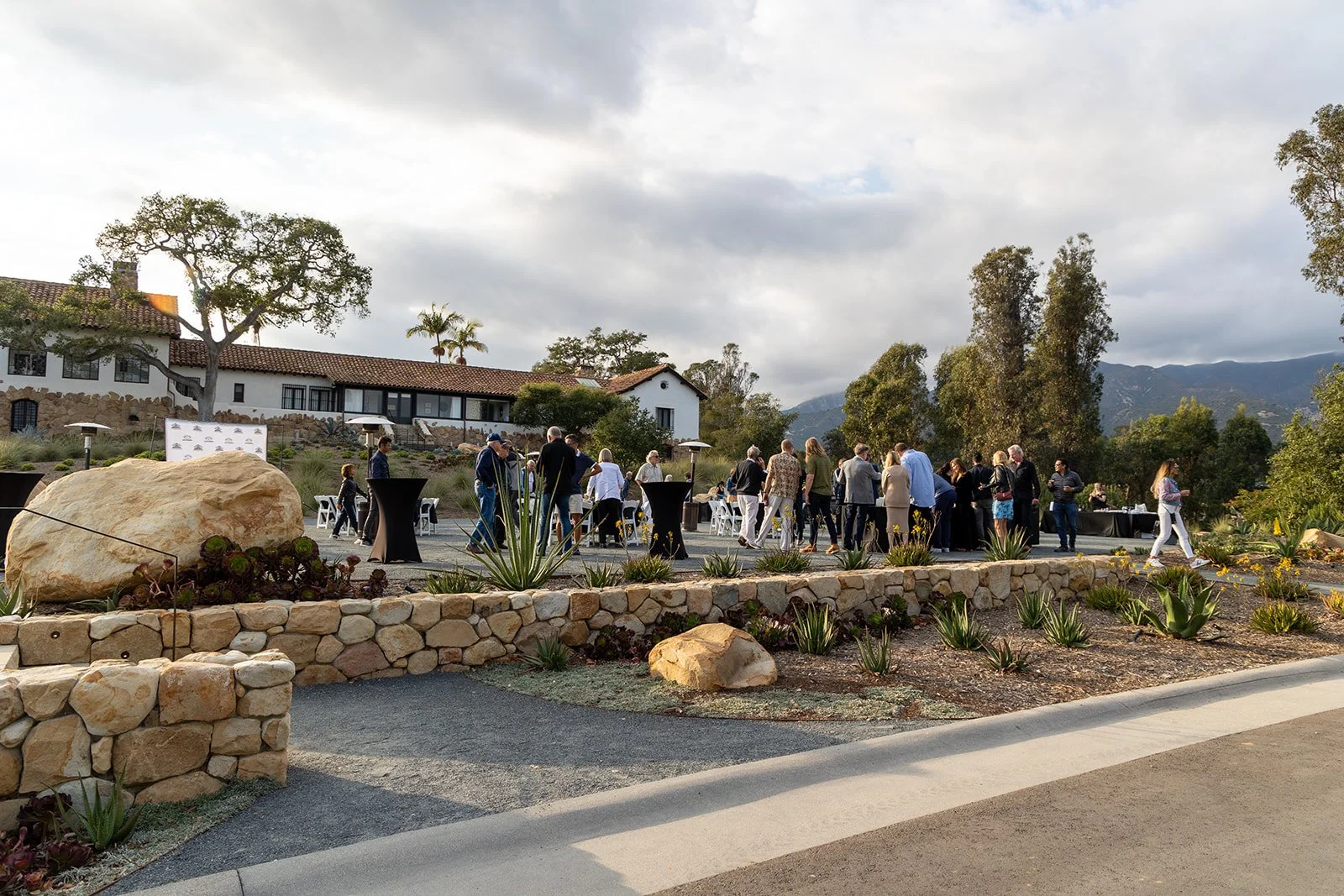People gathered outdoors for an event in a scenic setting with mountains, trees, and a building in the background.