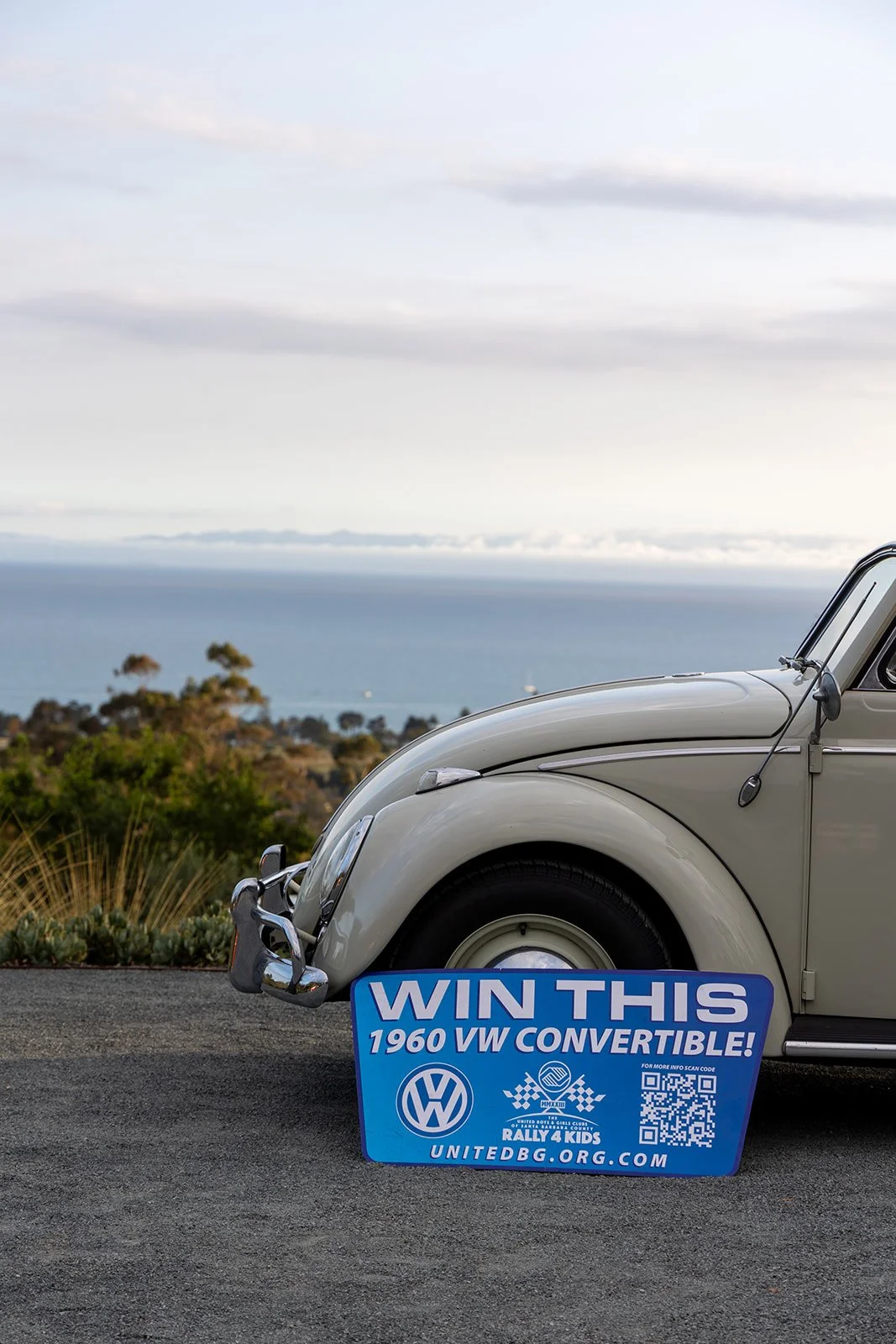 A cream-colored vintage Volkswagen Beetle parked near the coast with a blue sign in front that reads, 'Win This 1960 VW Convertible,' along with the Volkswagen logo, QR code, and website link for a children's charity.