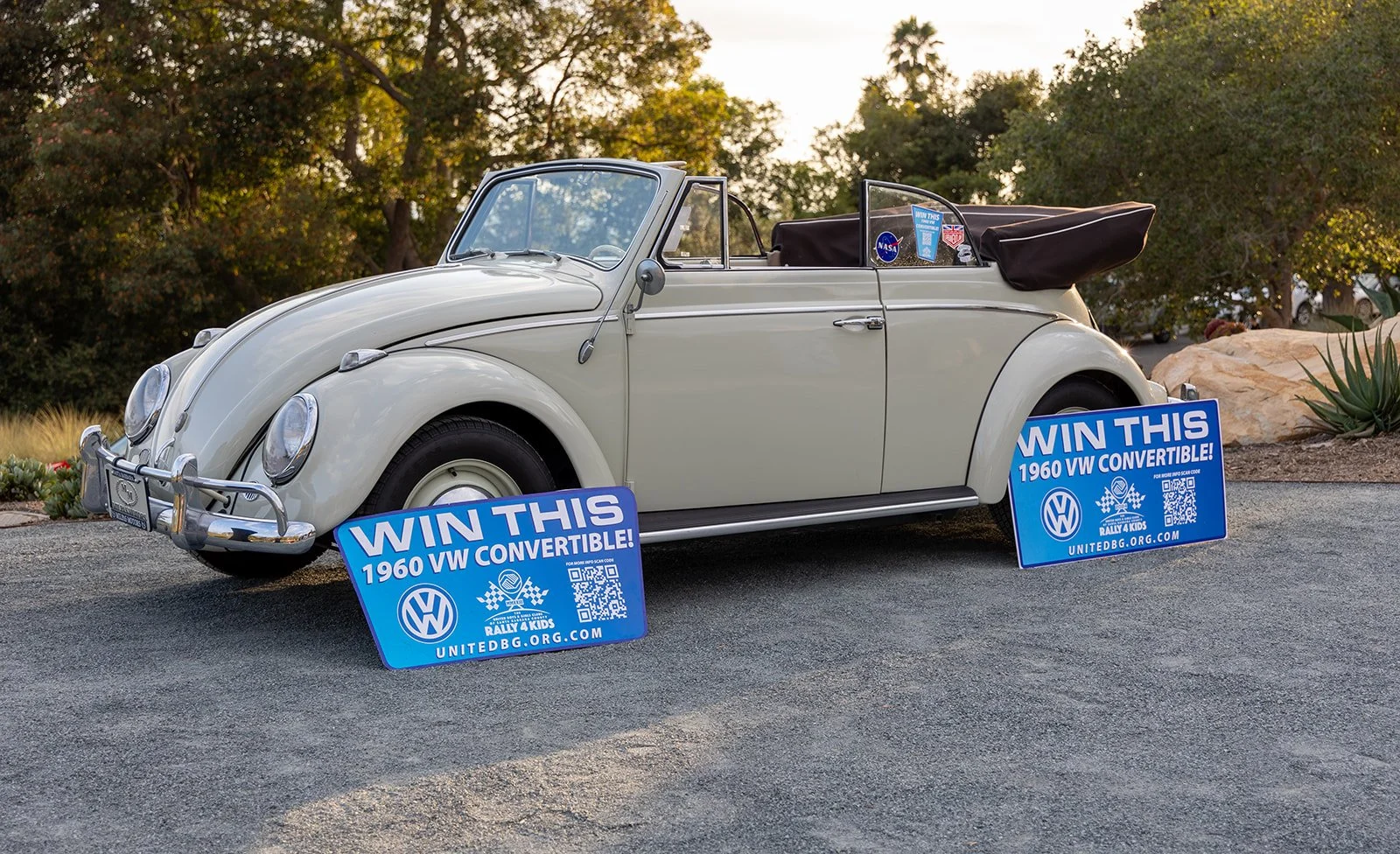 Vintage white Volkswagen convertible car parked outdoors with two blue signs in front promoting a contest to win a 1960 VW convertible, with organizers' website and a QR code. The car has a black soft top folded down, and the background includes tree
