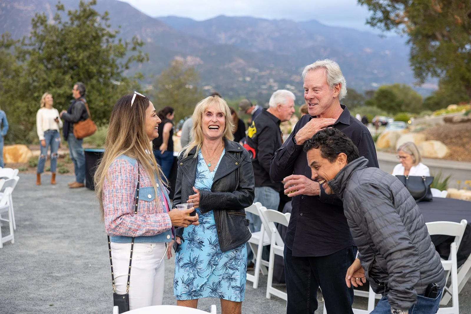 Group of people socializing outdoors at a gathering, smiling and laughing, with mountains in the background.