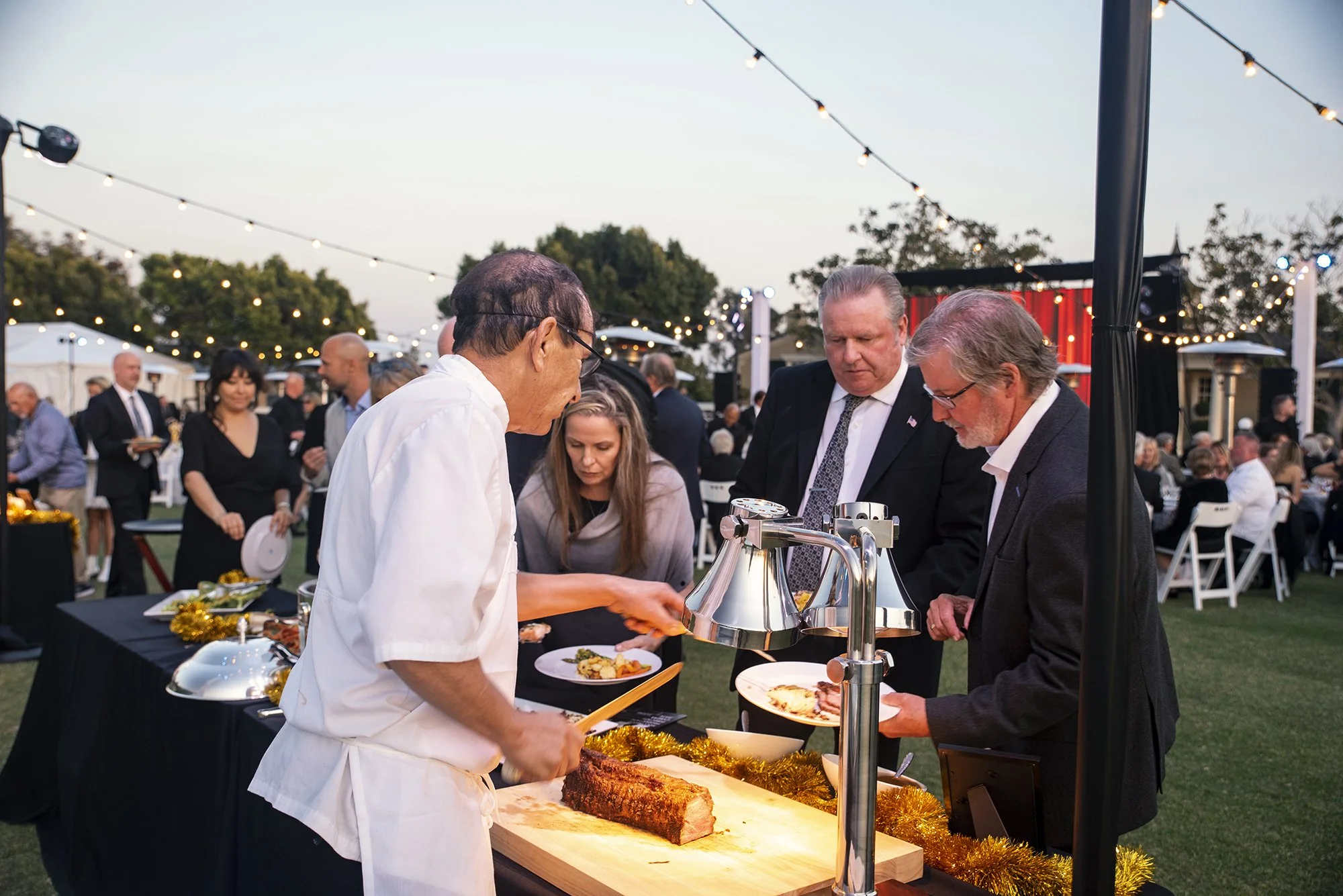 People at an outdoor evening event, some are serving and others are eating, with string lights overhead and tables in the background.