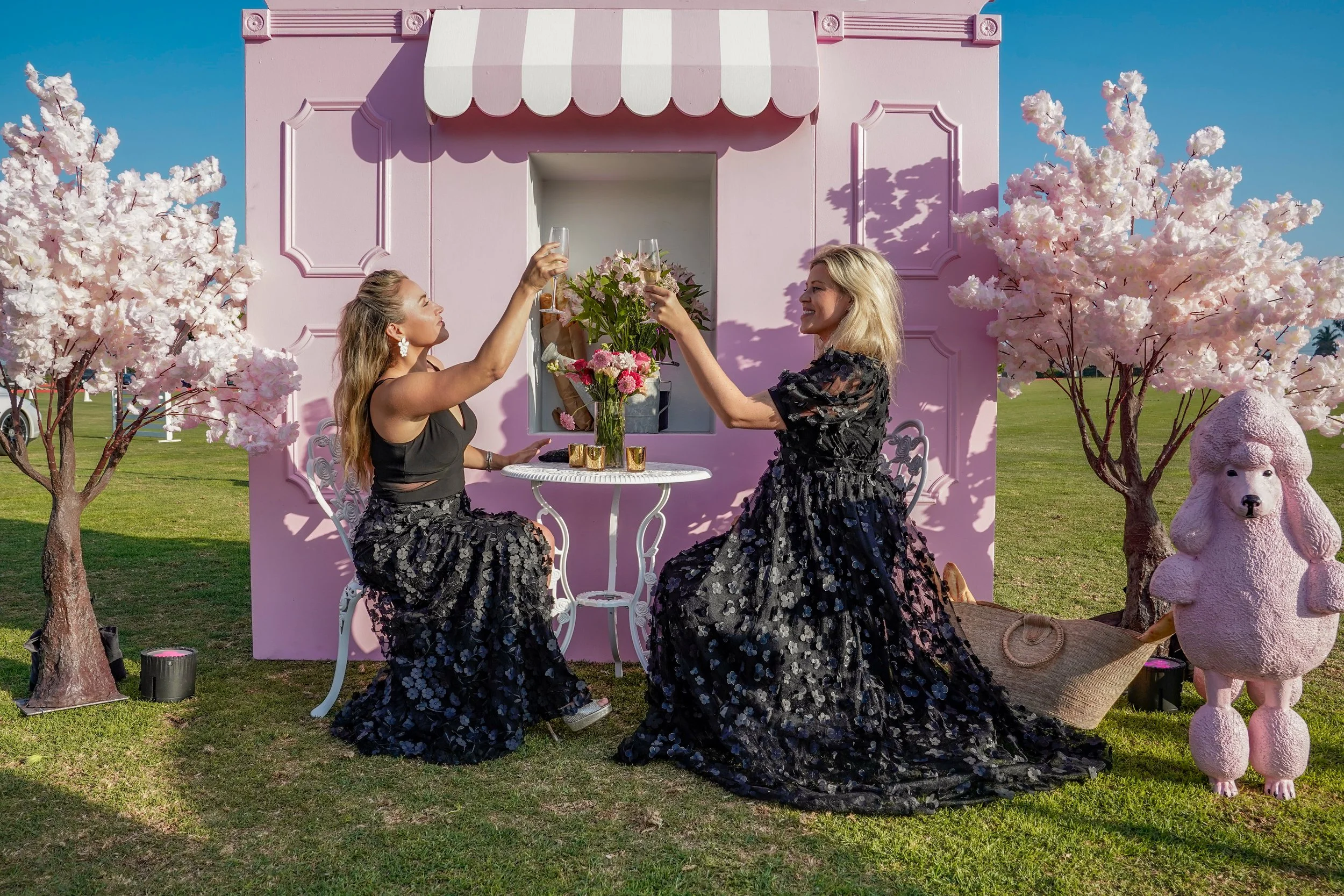 Two women in black floral skirts and black tops are having a picnic in a garden with pink trees and a pink building. They are raising glasses in a toast, sitting beside a table with flowers and a bouquet. A pink poodle statue is nearby.
