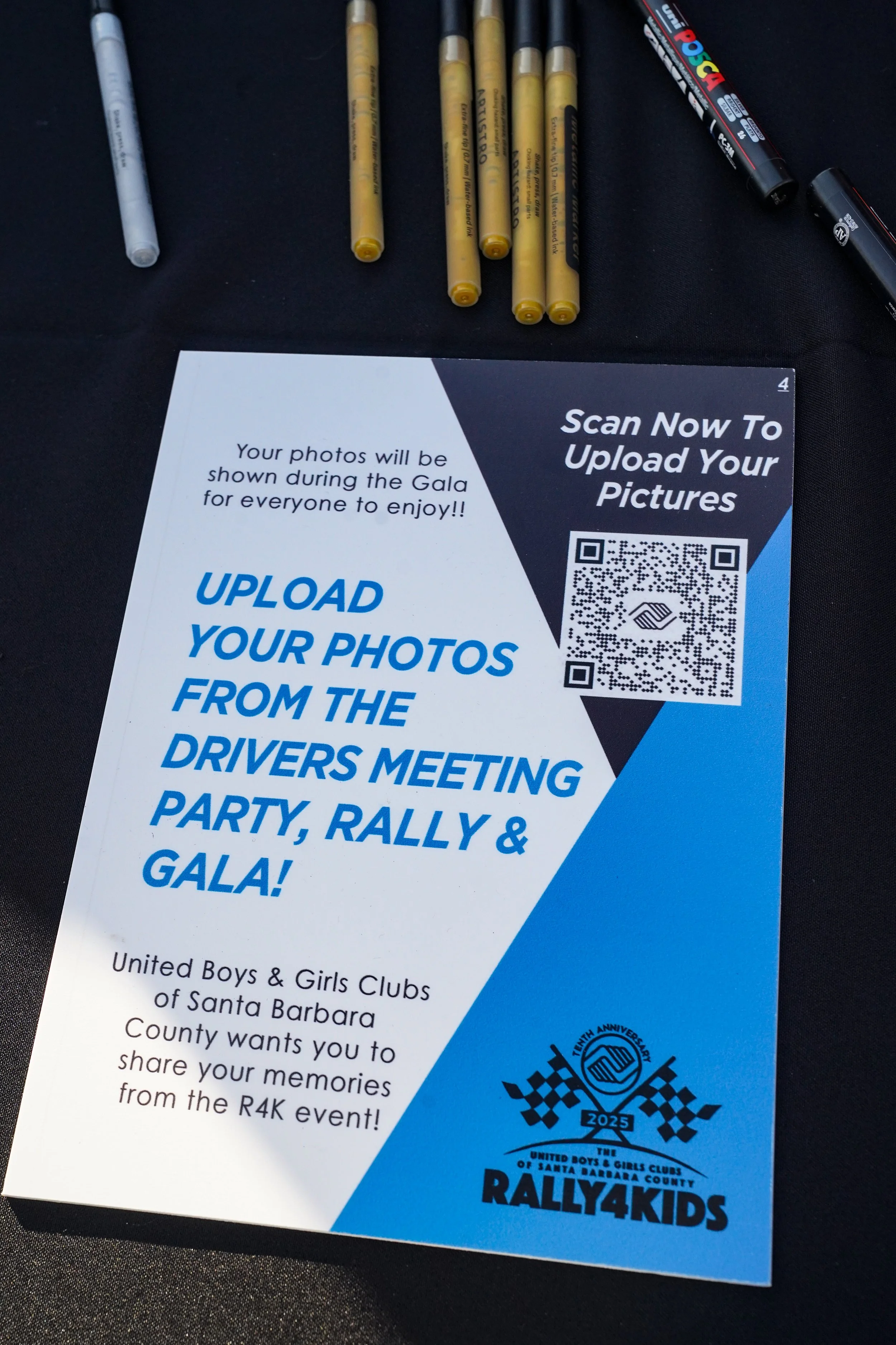A flyer on a black table encouraging people to upload photos from a drivers meeting, rally, and gala event via a QR code, with pens visible at the top of the image.