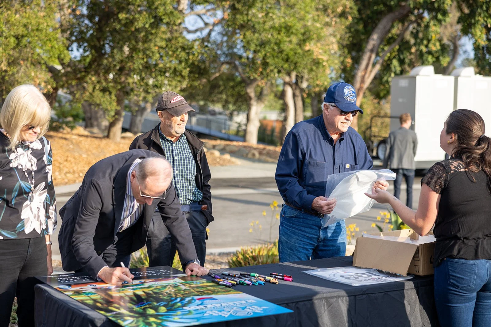 People attending an outdoor event, some are signing posters on a table with colorful markers, while others receive wrapped items. The setting has trees with fall foliage and a clear sunny sky.