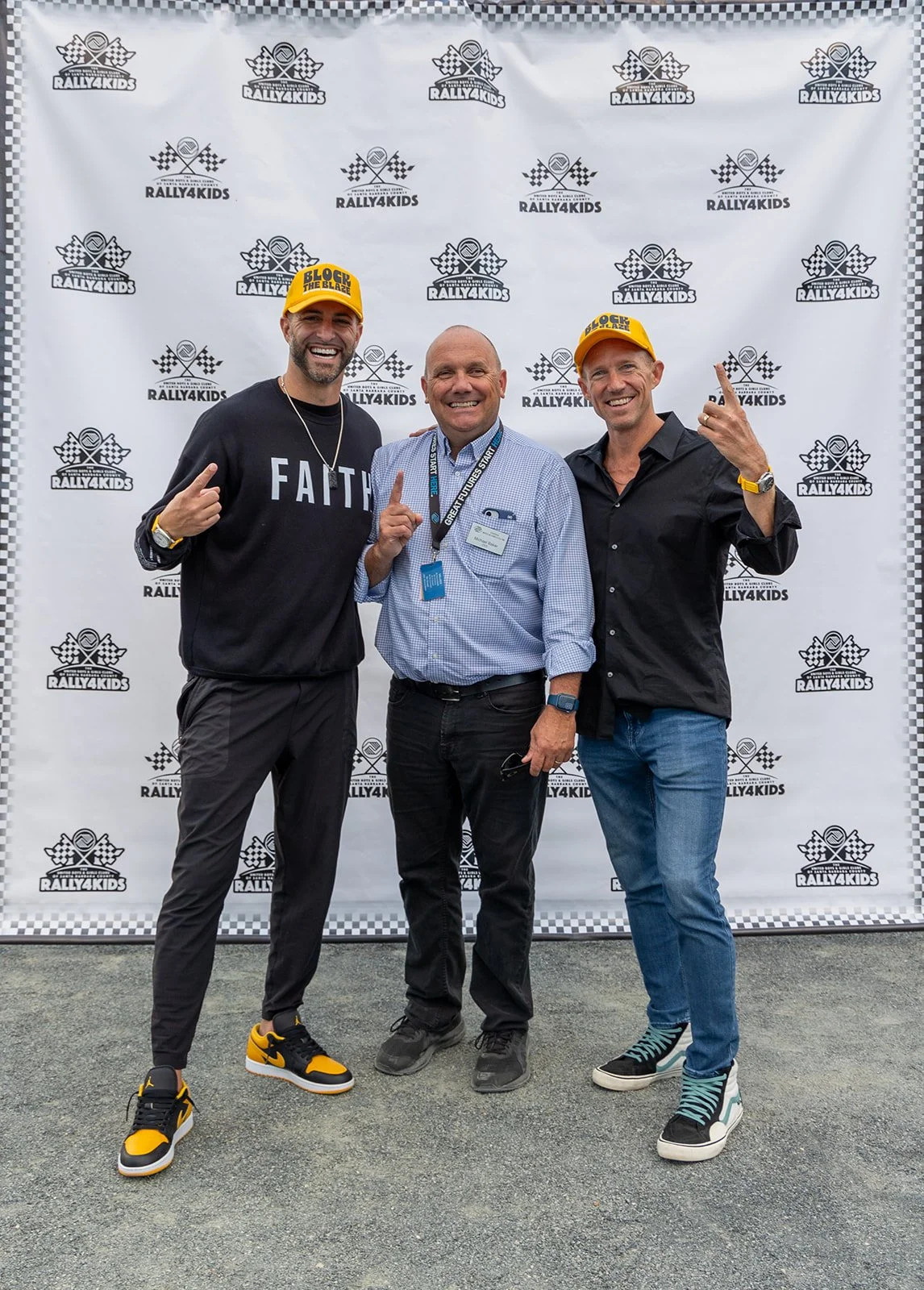 Three men standing in front of a Rally4Kids backdrop. Two men on either side are wearing yellow hats that say 'Block the Blaze' and casual clothing, making hand gestures. The man in the middle is wearing a blue shirt and a badge, smiling and holding 