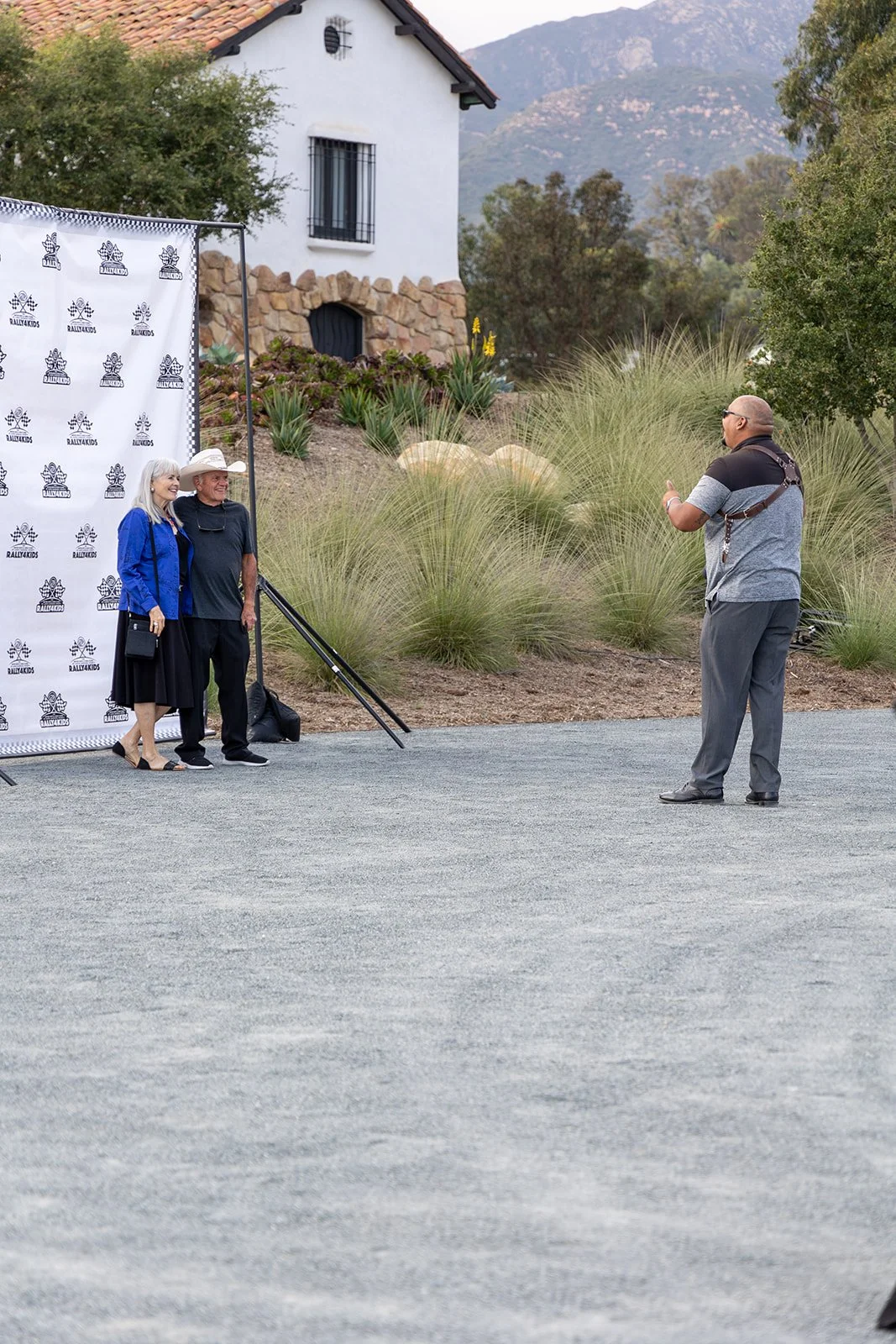 A photo of two elderly women posing for a picture with a man, outdoors in front of a branded backdrop, with a house and mountains in the background.