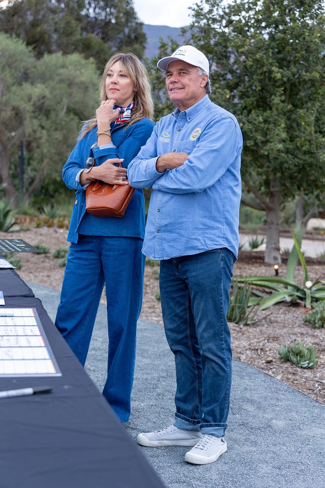 A man and woman stand outdoors, observing a display table. The woman has light hair, a blue jacket, and carries a brown purse. The man has gray hair, a white cap, a blue button-up shirt, and jeans. They are in a garden area with trees and plants.