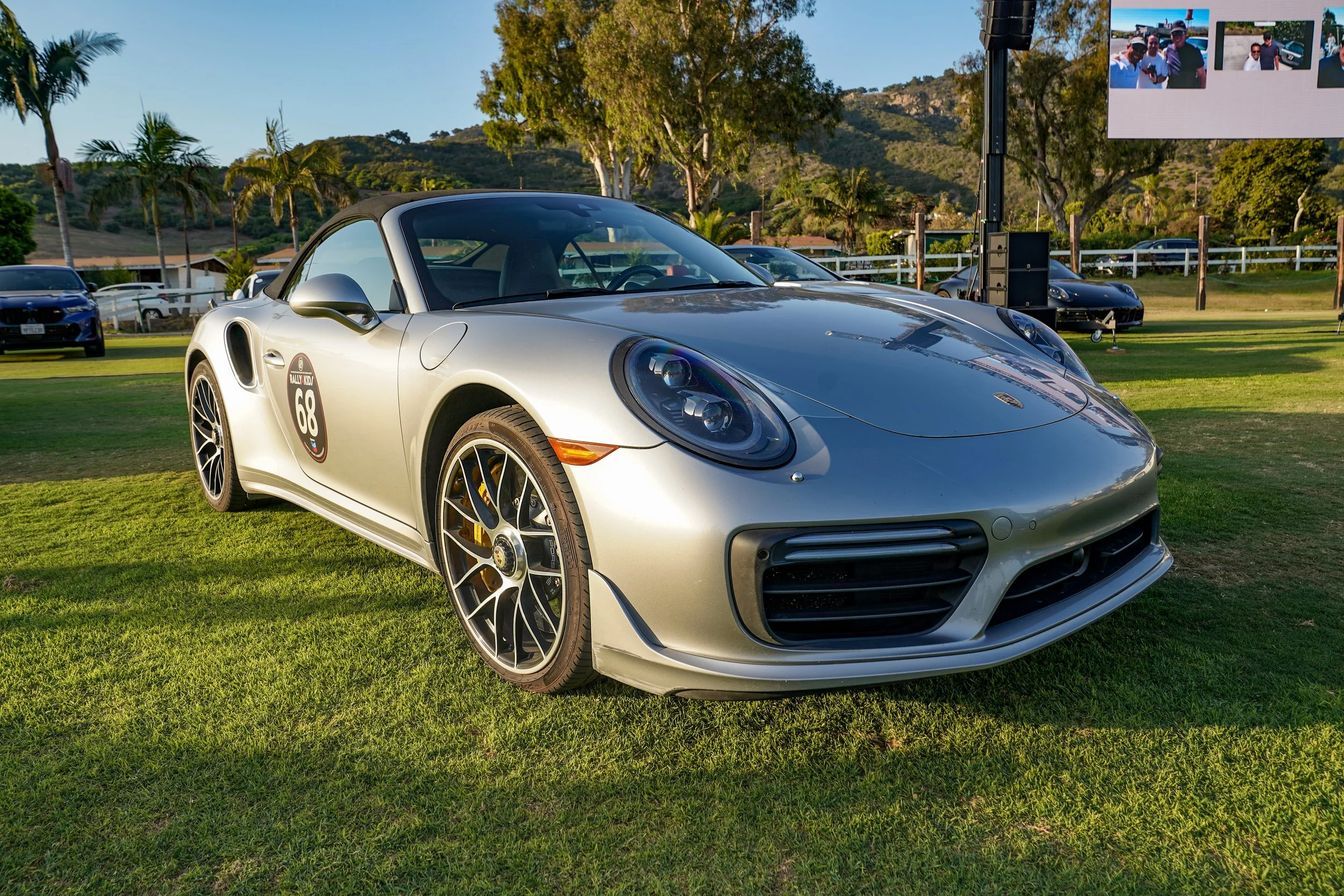 A silver Porsche 911 sports car with a rally number 68 decal on the door, parked on a grassy field during an outdoor event.