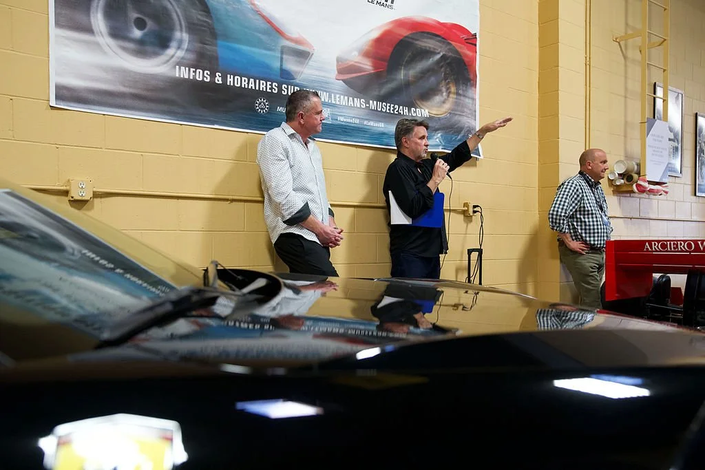 Three men speaking at an indoor event with a large car advertisement banner in the background, yellow brick wall, and a black car in the foreground.