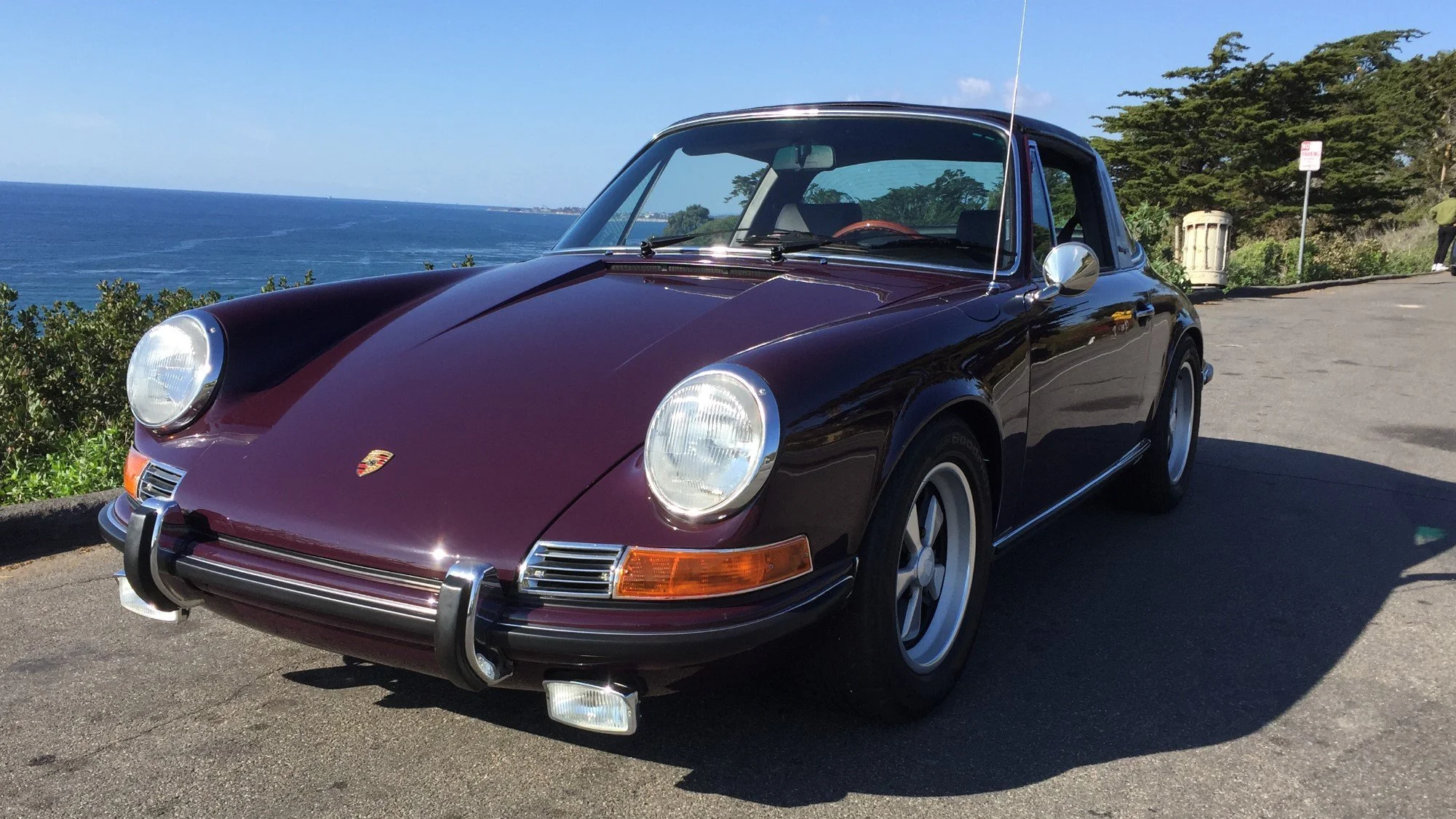 A burgundy vintage Porsche 911 parked near the coast with ocean and sky in the background.