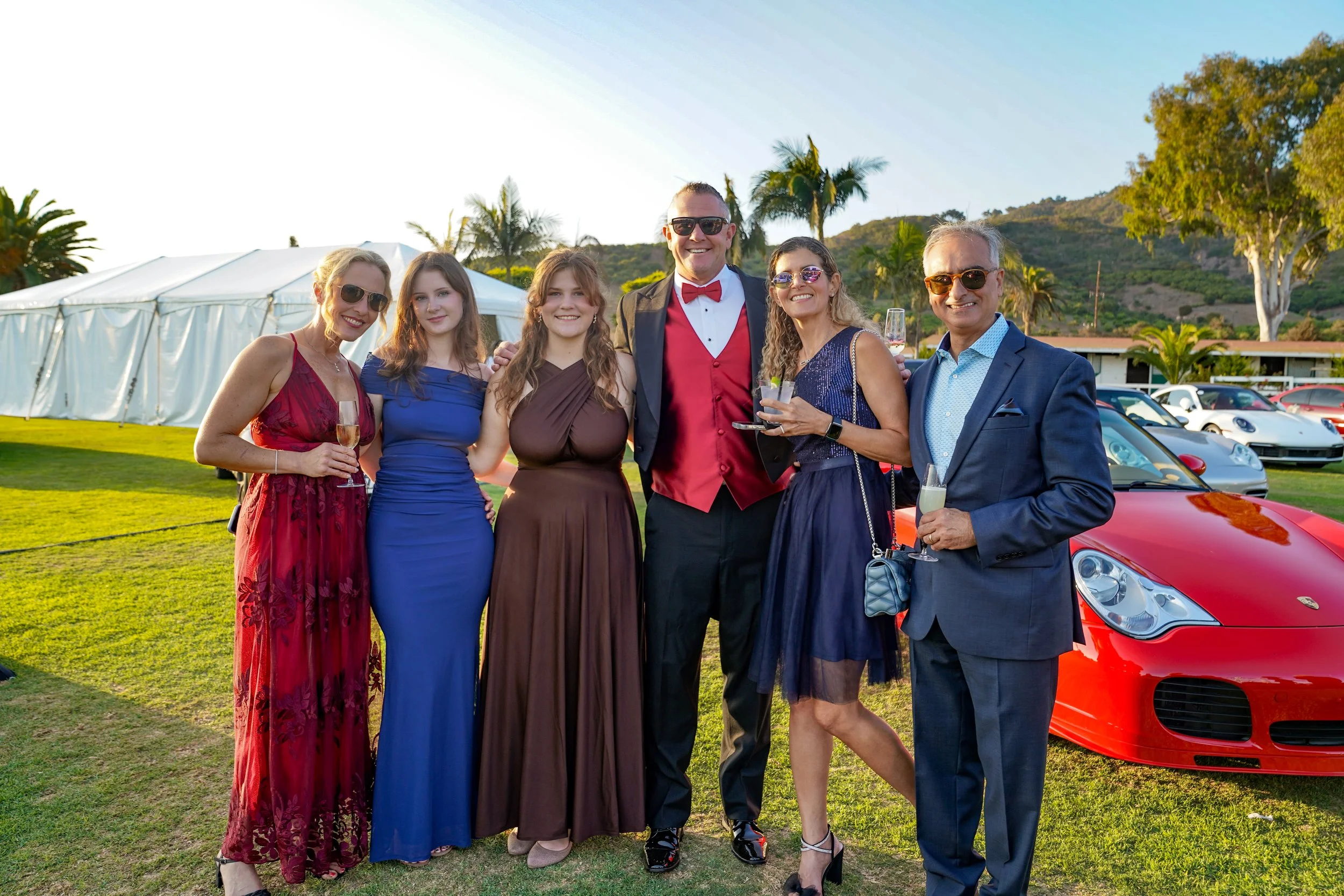 Group of seven adults dressed in formal attire at an outdoor event, some holding champagne glasses, with a red sports car and a white marquee tent in the background.