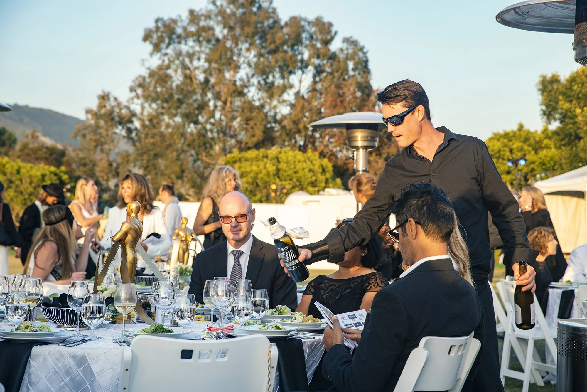 People attending an outdoor formal event, seated at a table with white tablecloths, wine glasses, and decorated with gold award statues, with a waiter serving drinks to guests.