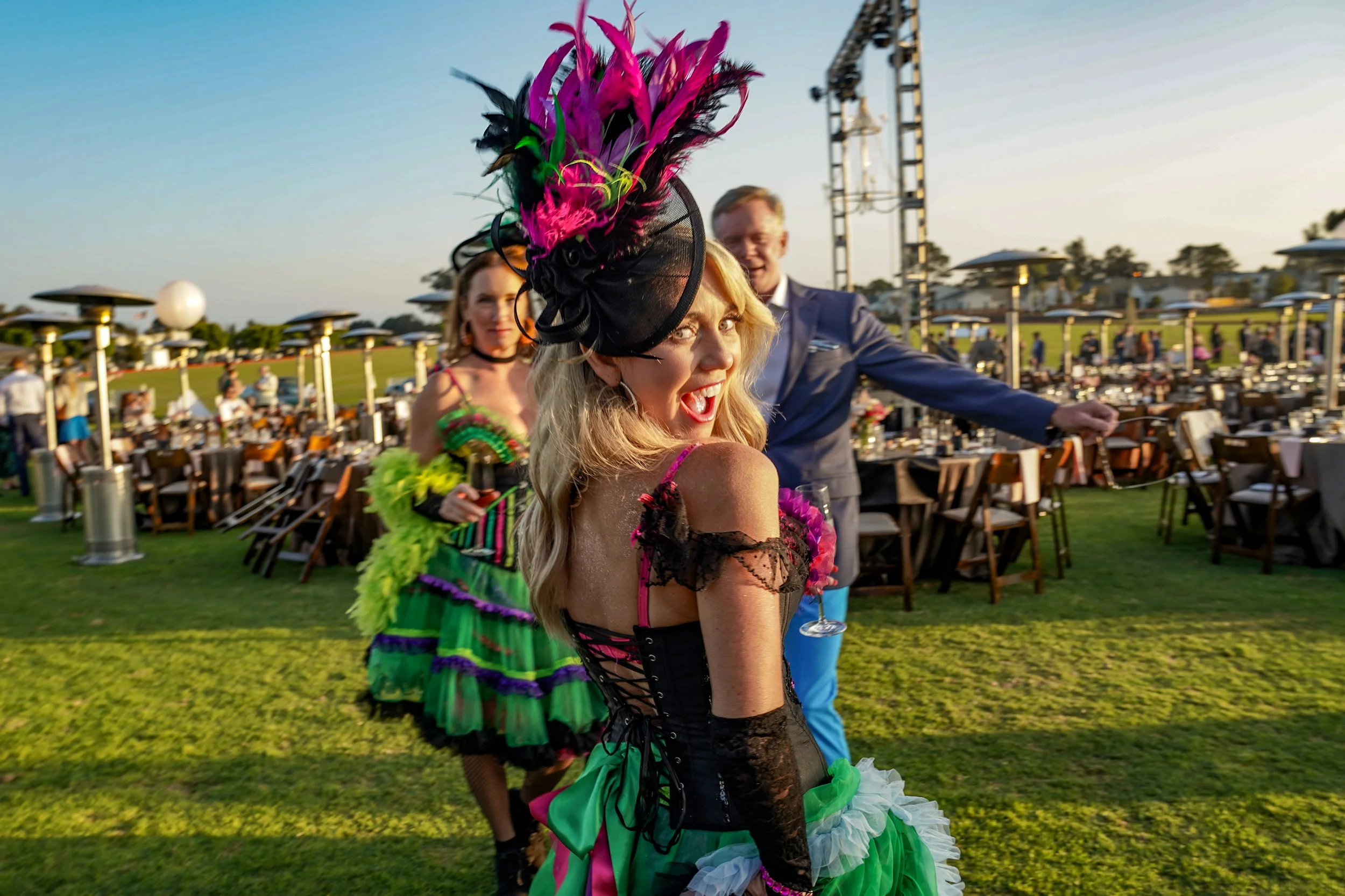 Woman in colorful, elaborate costume with black and pink feathered hat posing at outdoor event during sunset, with others in vibrant costumes and banquet tables in background.