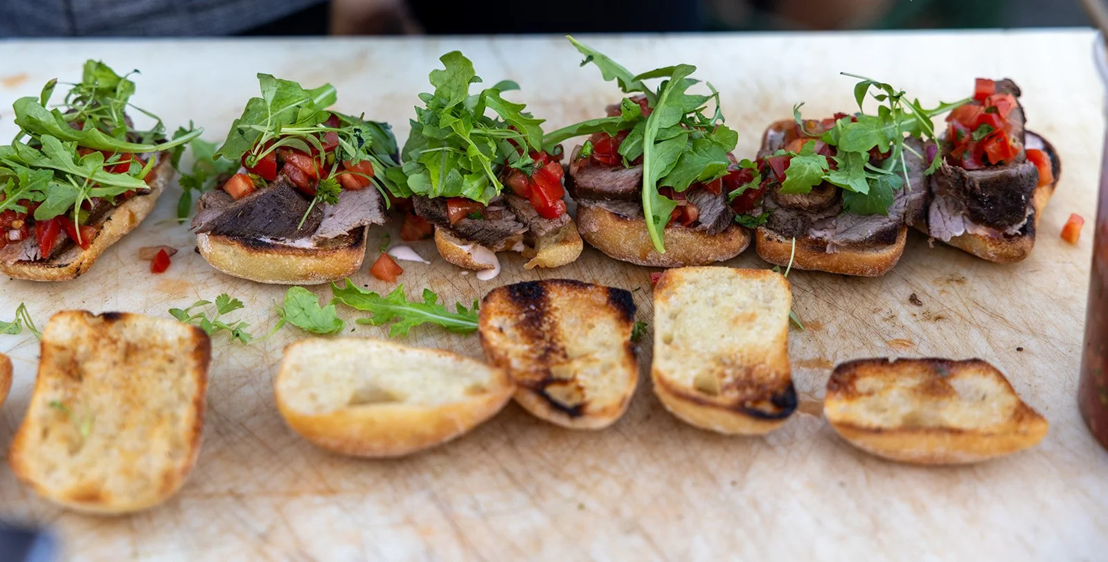 Open-faced sandwiches with sliced roast beef, chopped red peppers, and arugula on toasted bread, with toasted bread pieces on the side on a wooden cutting board.