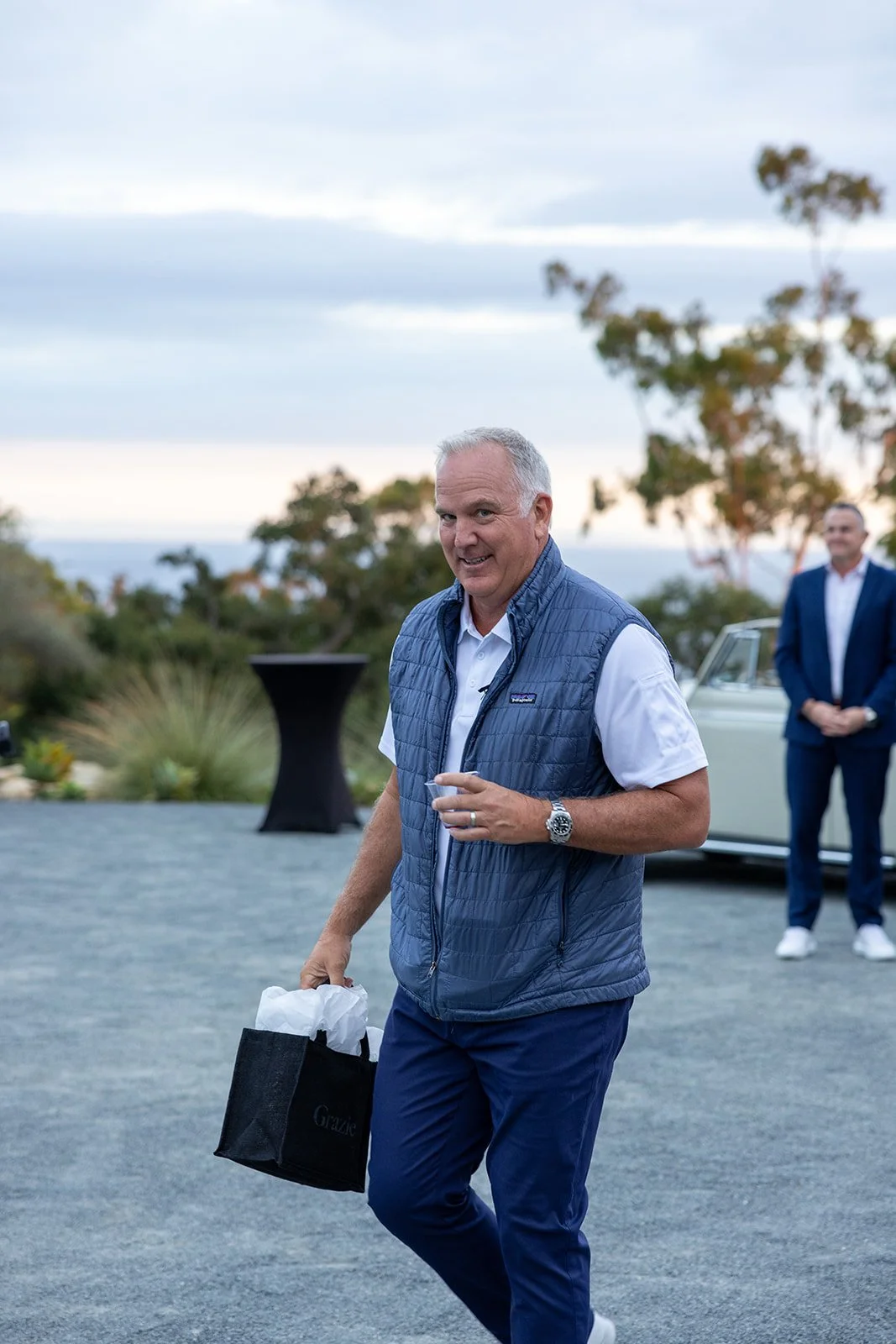 Man holding a black gift bag with tissue paper, smiling outdoors during evening, with another man in a suit and a vintage car in background.
