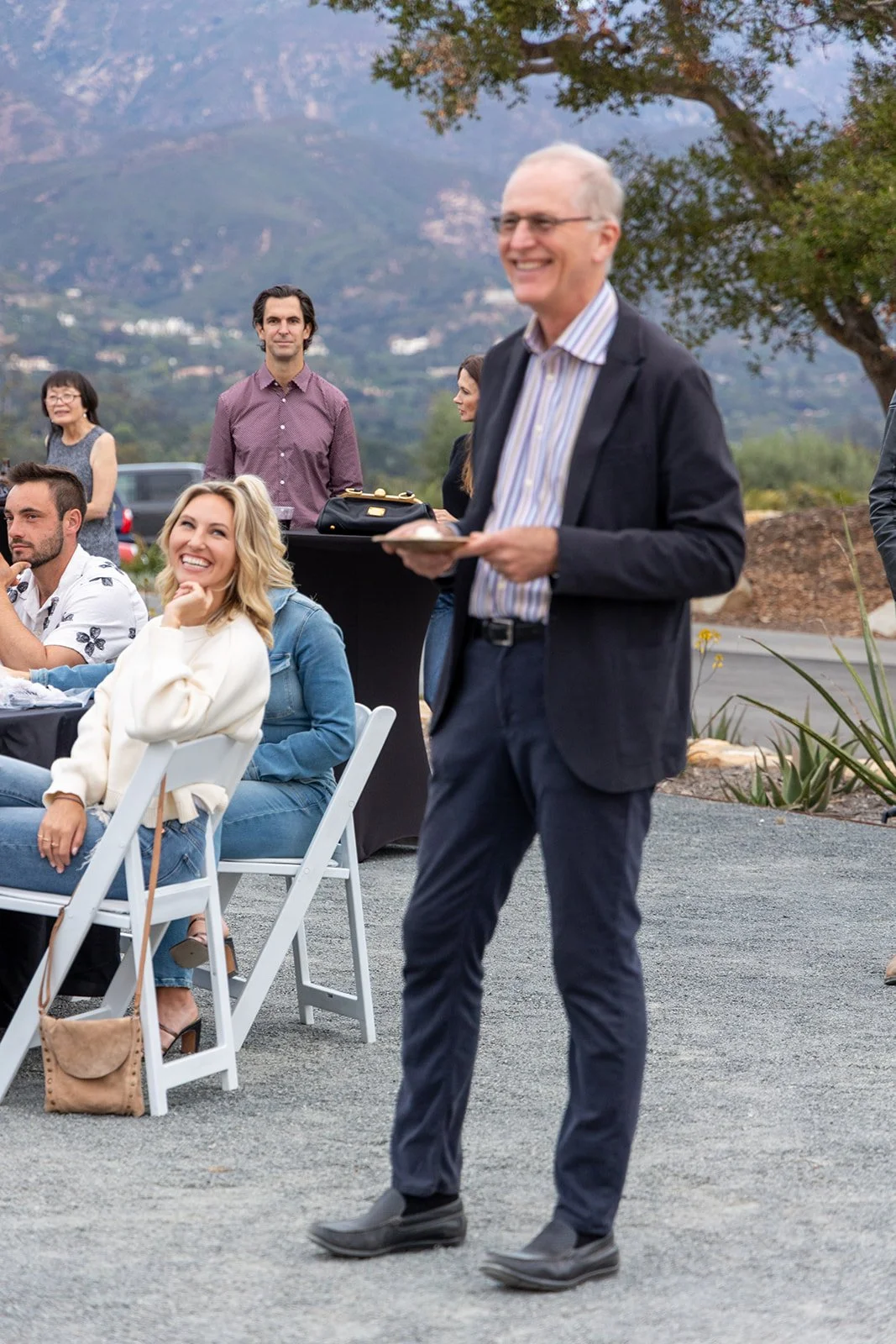 Man in suit standing and smiling at an outdoor social gathering with seated and standing people, mountains in the background.