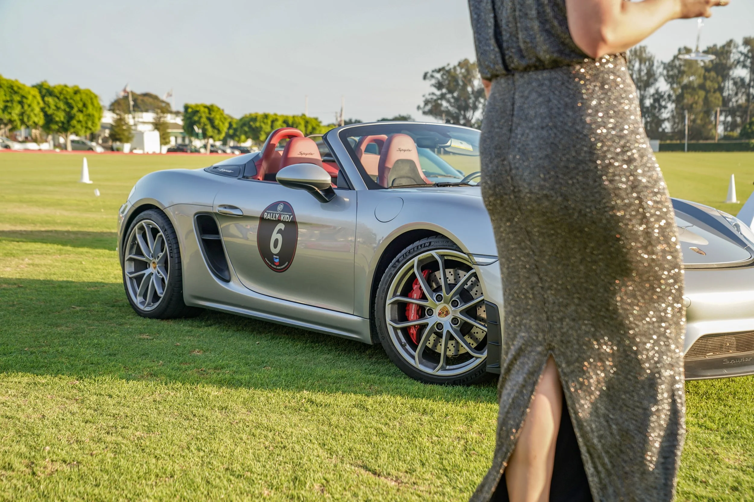 Silver Porsche sports car with a race number 6 sticker on the side, parked on a grassy field, with a woman in a shiny gold dress standing nearby, partially visible in the foreground.