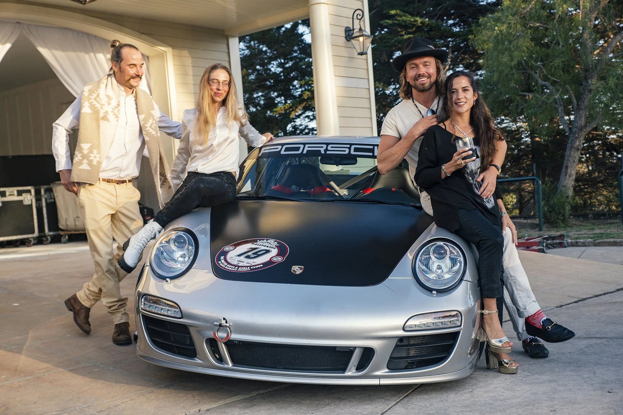 Group of four people standing and sitting around a silver Porsche race car with black hood, parked outside a house with a garage and trees in the background. Two men and two women, dressed in casual and stylish clothing, smiling on a sunny day.