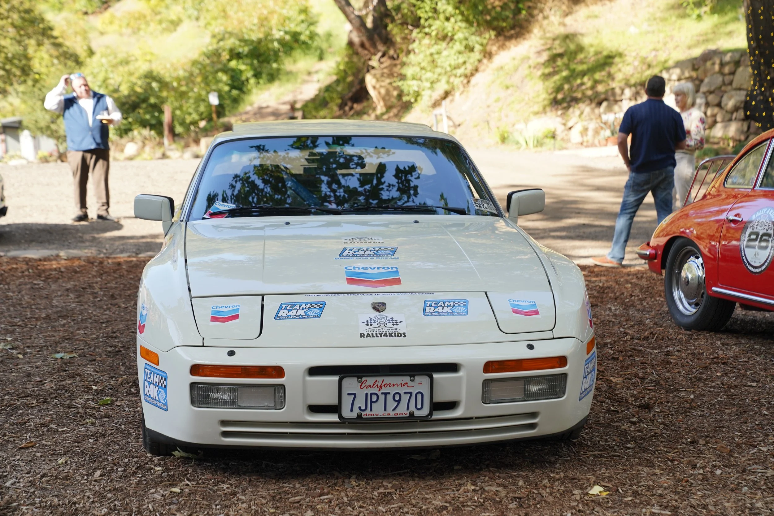 Front view of a white Porsche with racing stickers, parked on gravel, with people in the background at an outdoor car event.