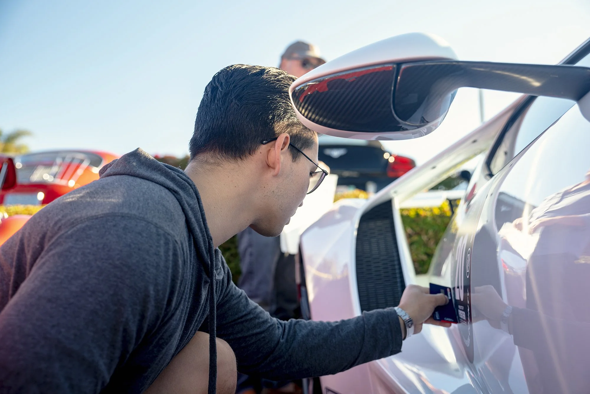 A man with dark hair, glasses, and a gray hoodie is kneeling beside a white sports car, attaching a sticker to its side. Another person stands in the background, and there are other sports cars outdoors on a sunny day.