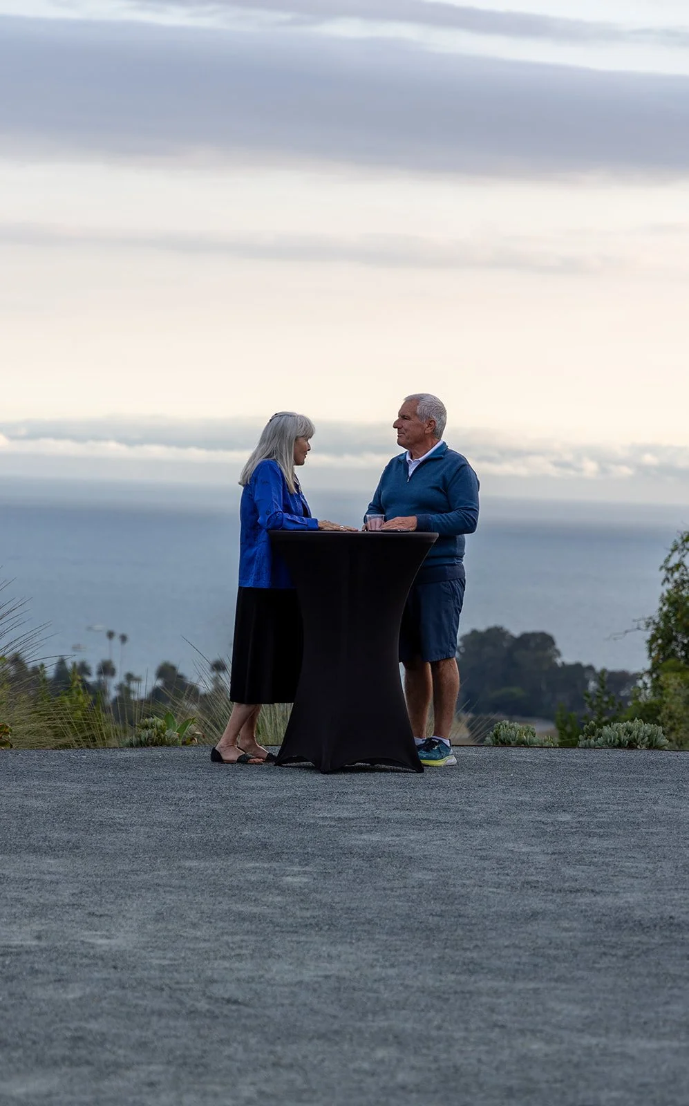 A senior couple standing at a tall table outdoors near the coast during cloudy weather, with the ocean and trees in the background.