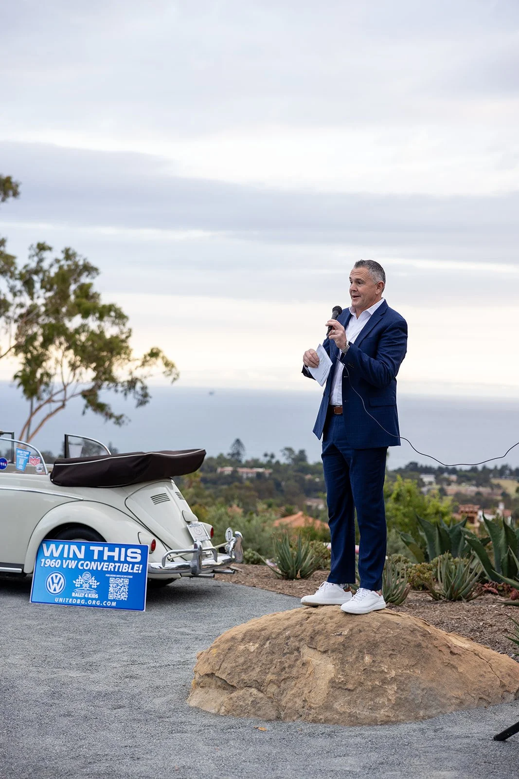 Man in a suit speaking into a microphone outdoors, next to a vintage Volkswagen convertible car with a sign that says "Win This 1960 VW Convertible!"