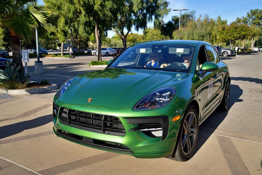 A green Porsche Macan parked in a parking lot with trees and other vehicles in the background.