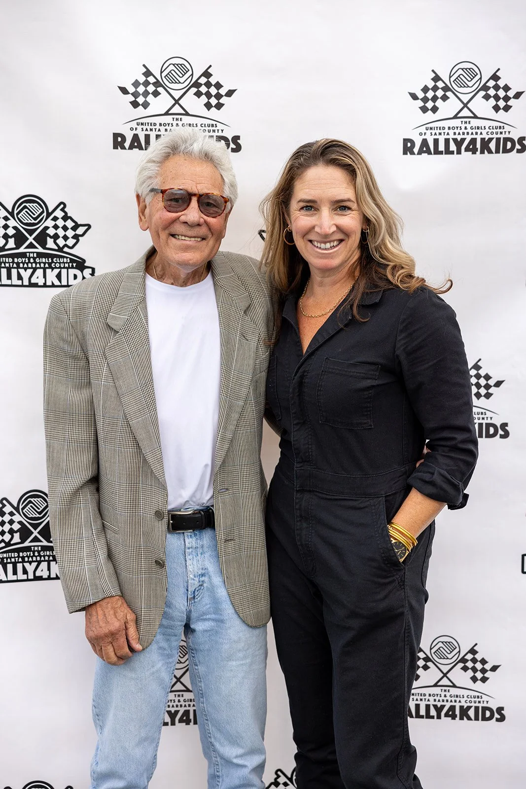 An older man with white hair, glasses, in a plaid blazer and jeans, standing next to a woman with long wavy hair in a black jumpsuit, both smiling, at a rally event with a backdrop featuring the logo for Rally4Kids.