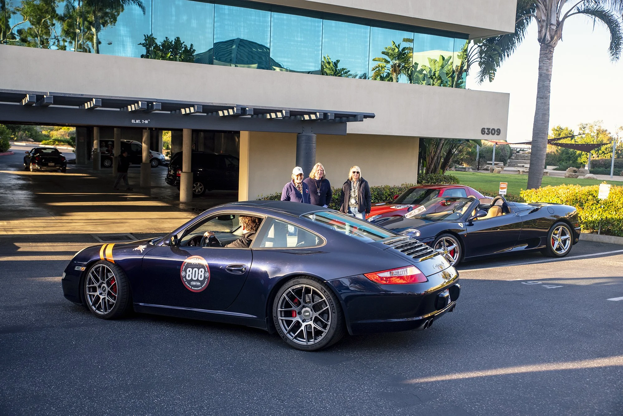 Three women standing near a black Porsche sports car and two other sports cars parked outside a modern building with glass windows and palm trees.