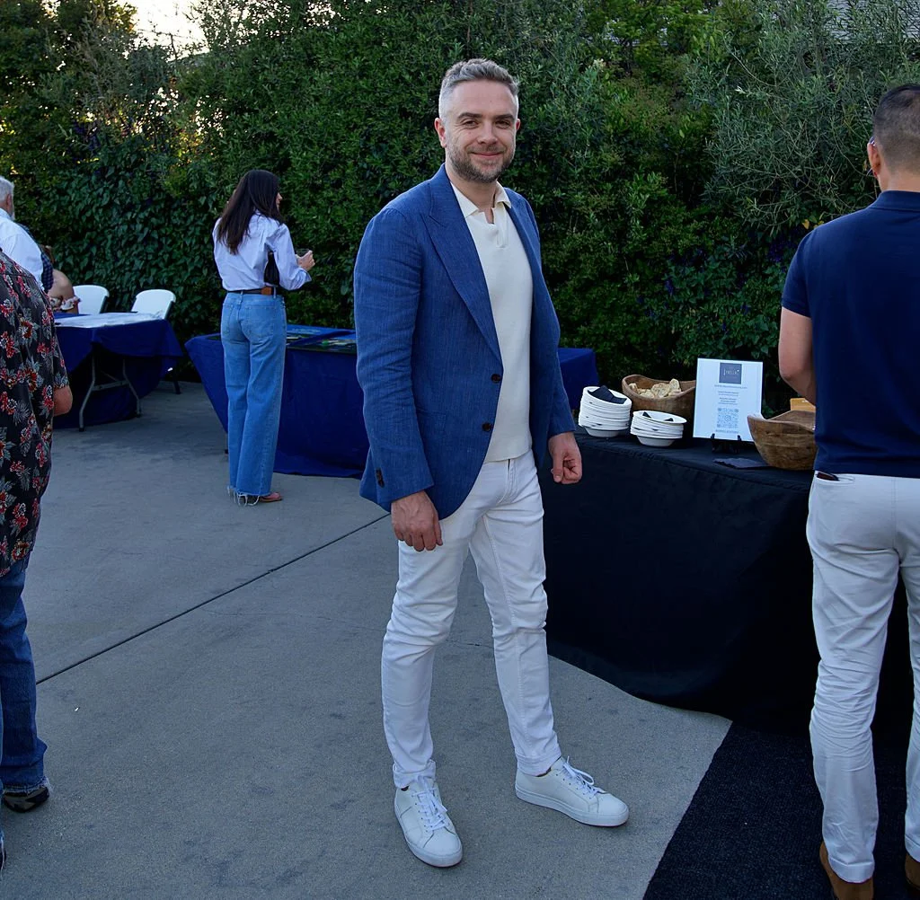A man in a blue blazer and white pants standing outdoors at a social gathering, with a buffet table behind him.