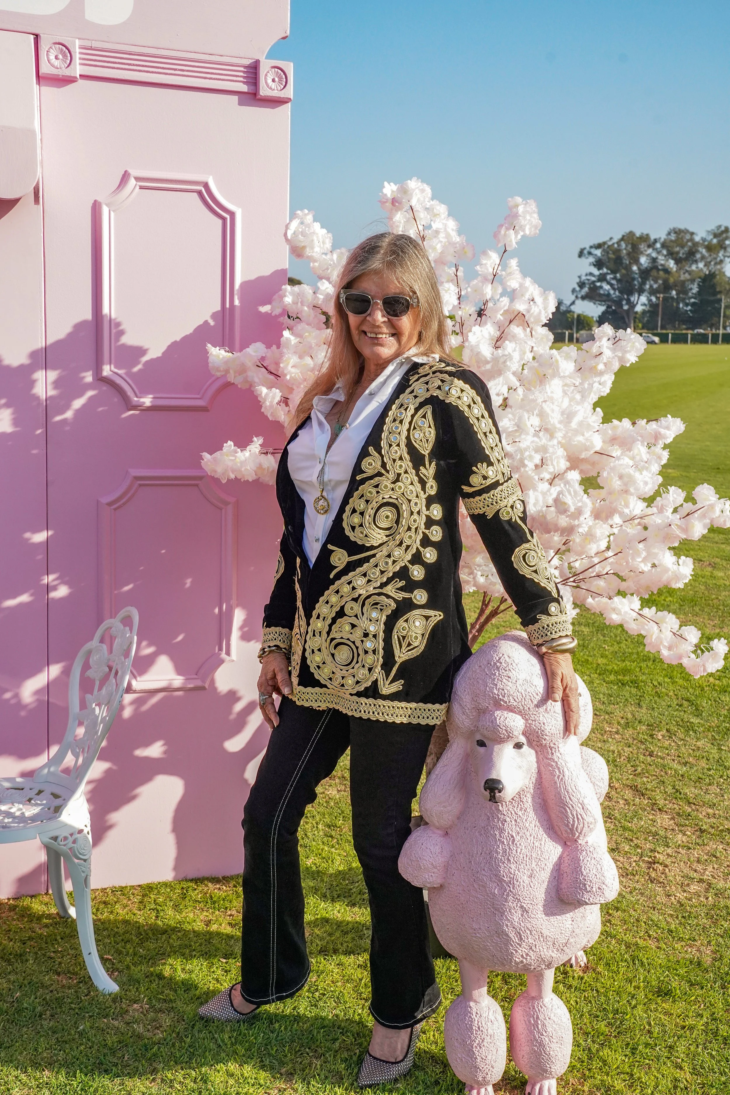 A smiling woman with blond hair, sunglasses, and a black and gold embroidered jacket standing outside. She's holding a pink poodle-shaped sculpture and is positioned next to a pink door, a white chair, a pink flowering tree, and a grassy field under 