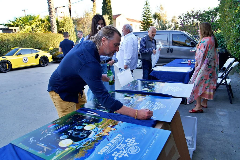 People gathering outdoors, signing posters on a table, with a yellow sports car and a gray van in the background.