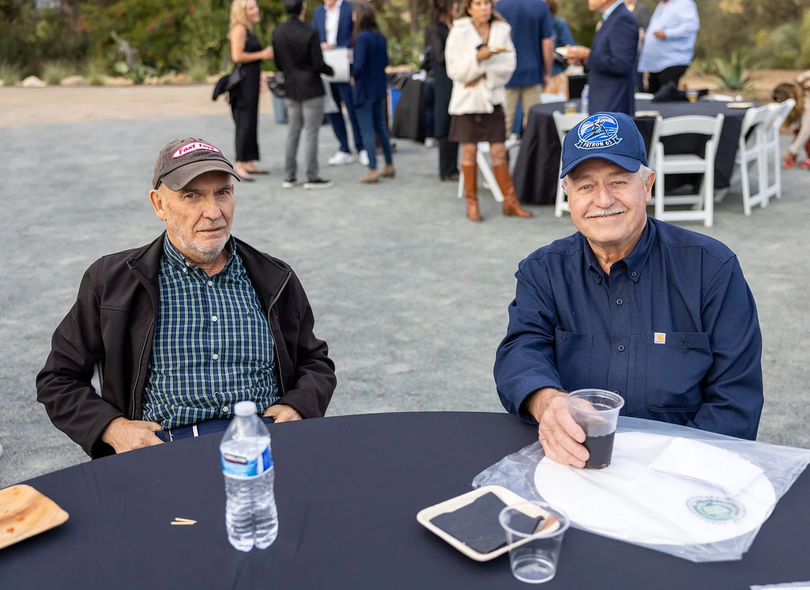 Two older men sitting at a round table outdoors at a social event. The man on the left is wearing a dark jacket and a baseball cap, while the man on the right is smiling and wearing a dark blue shirt with a Carhartt logo and a blue cap with a logo, h