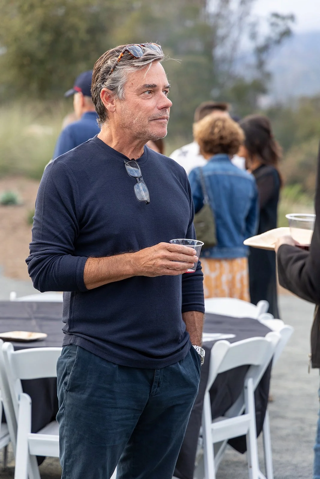 Man with sunglasses on his head holding a clear plastic cup, attending outdoor gathering with people in the background.